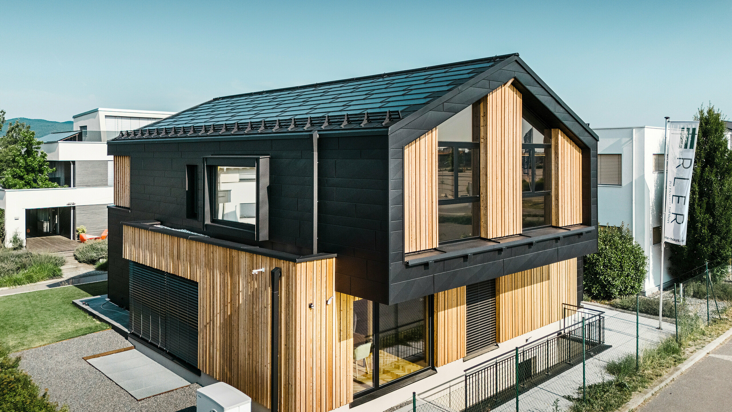 Parte anteriore della casa modello Autark nel centro di case prefabbricate Blaue Lagune, Vösendorf (AT). Il tetto è ricoperto da tegola fotovoltaica PREFA nera e tegola R.16 con verniciatura P.10 di alta qualità. La facciata è costituita da una combinazione di elementi verticali in legno e PREFA Doga.X in P.10 nero, che conferisce all'edificio un carattere moderno e sostenibile. Sul bordo destro dell'immagine sventola la bandiera della Erler Bau GmbH.