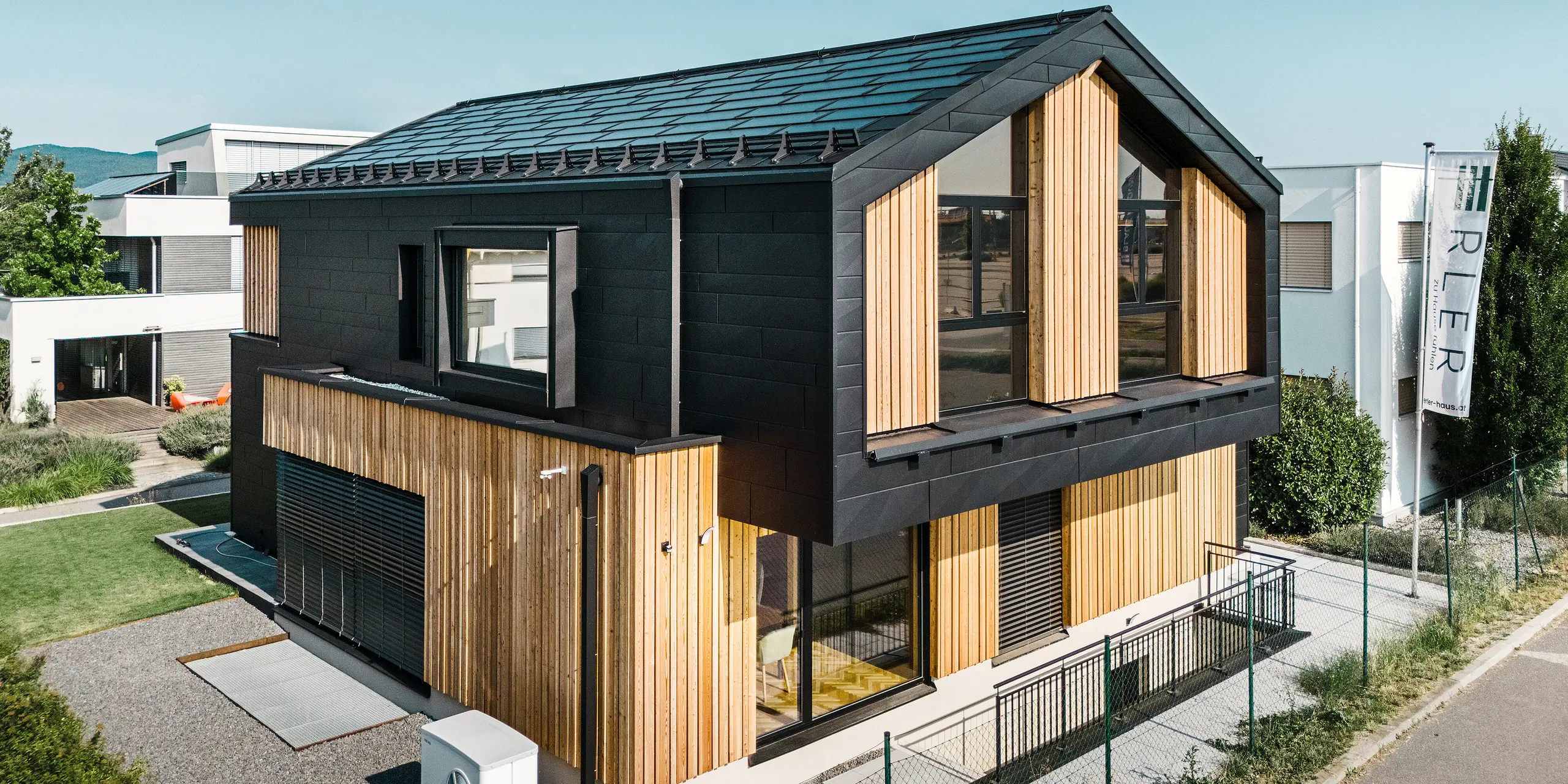 Parte anteriore della casa modello Autark nel centro di case prefabbricate Blaue Lagune, Vösendorf (AT). Il tetto è ricoperto da tegola fotovoltaica PREFA nera e tegola R.16 con verniciatura P.10 di alta qualità. La facciata è costituita da una combinazione di elementi verticali in legno e PREFA Doga.X in P.10 nero, che conferisce all'edificio un carattere moderno e sostenibile. Sul bordo destro dell'immagine sventola la bandiera della Erler Bau GmbH.