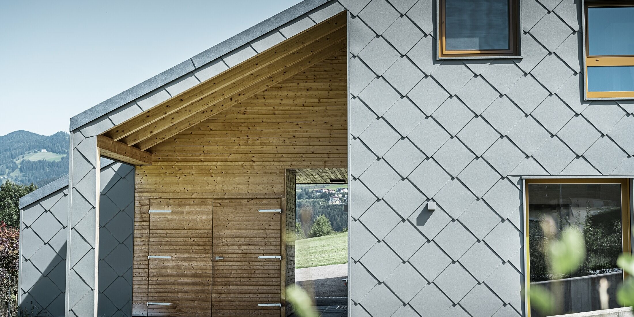 L'ingresso coperto è realizzato con un rivestimento orizzontale in legno, mentre il resto della facciata è rivestito con la scaglia per facciata PREFA grande in alluminio grigio chiaro.