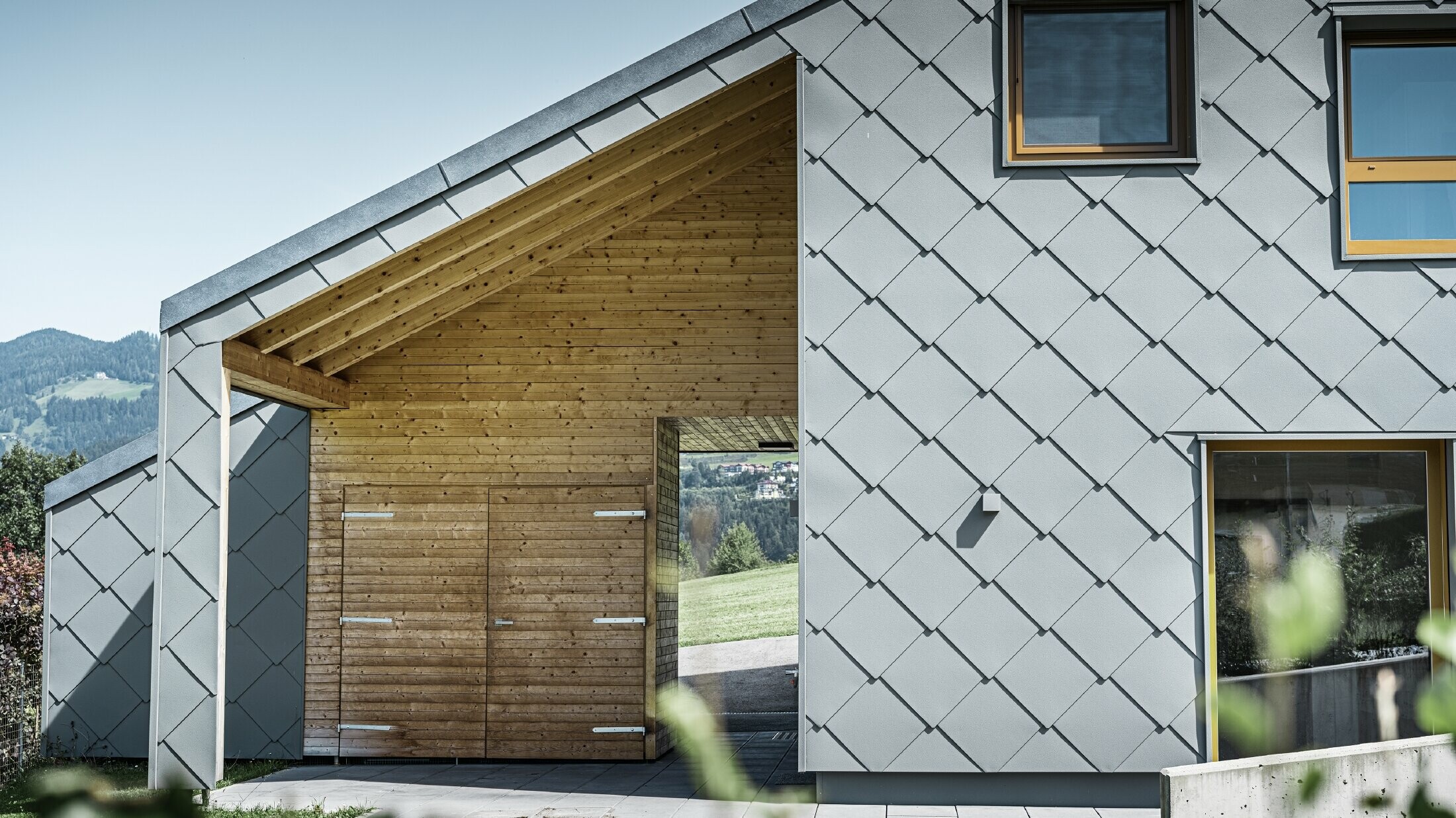 L'ingresso coperto è realizzato con un rivestimento orizzontale in legno, mentre il resto della facciata è rivestito con la scaglia per facciata PREFA grande in alluminio grigio chiaro.
