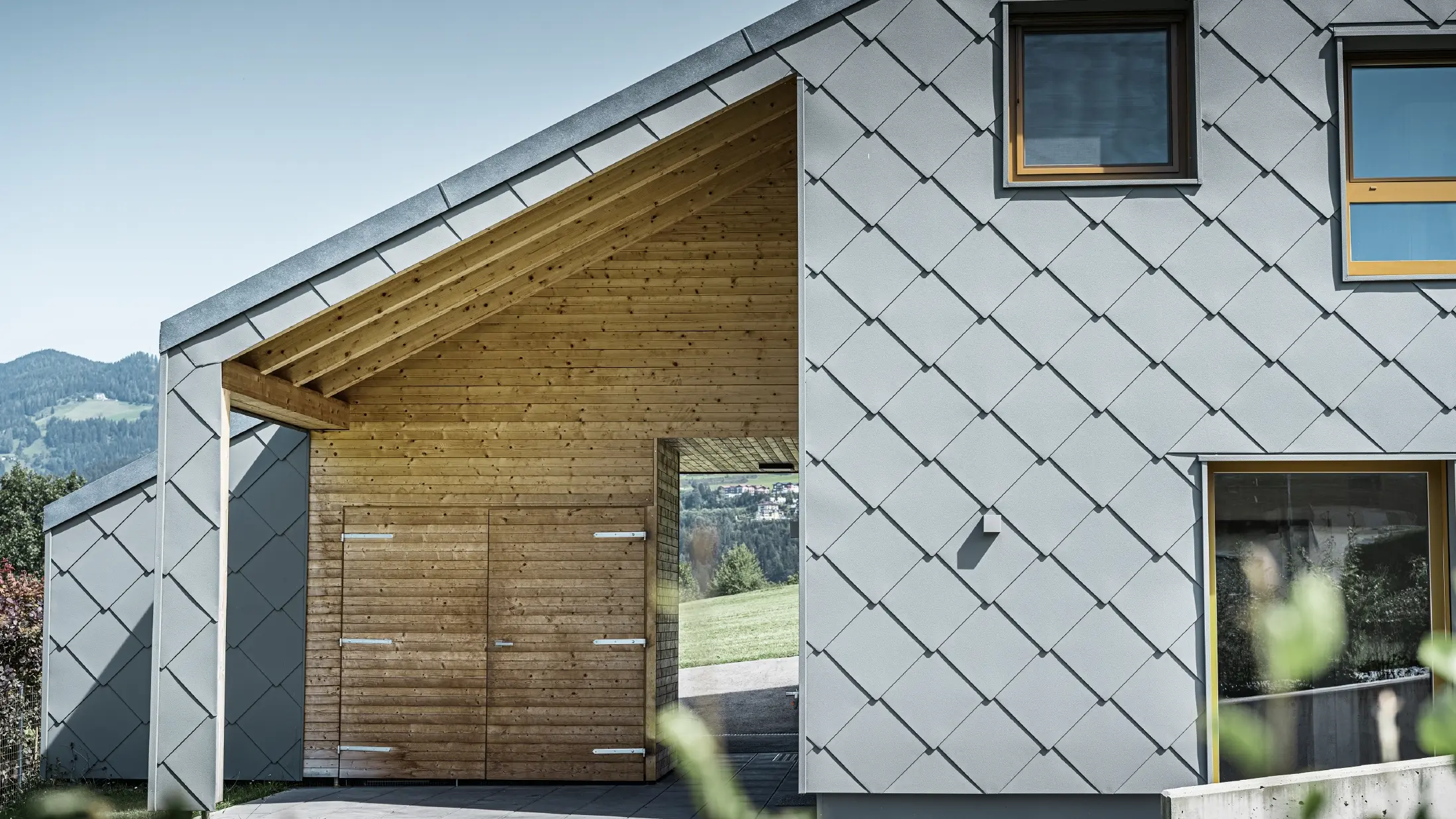 Der überdachte Eingangsbereich ist mit waagrechter Holzverkleidung gemacht, der Rest der Fassade ist mit der großen PREFA Wandraute aus Aluminium in Hellgrau verkleidet
