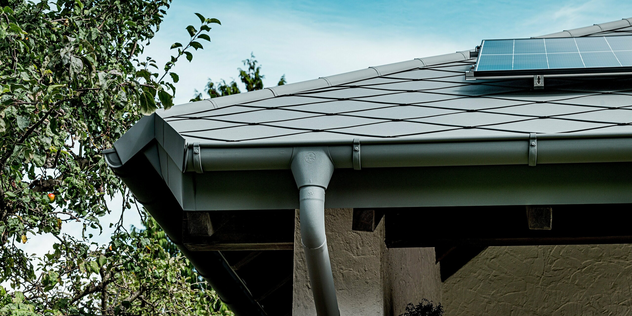 Das Bild zeigt eine Nahaufnahme der Traufe eines Einfamilienhauses, eingedeckt mit PREFA Dachrauten 44 × 44 in P.10 Hellgrau. Die markante Rautenstruktur und die saubere Linienführung betonen die präzise Verarbeitung des Aluminiumdachs. Im Vordergrund ist die robuste PREFA Hängerinne mit passendem Ablaufrohr zu sehen, die harmonisch in das System integriert ist. Eine Ecke der Photovoltaikanlage ragt ins Bild und verdeutlicht die ideale Kombination aus langlebigem PREFA Dach und moderner Solartechnik.