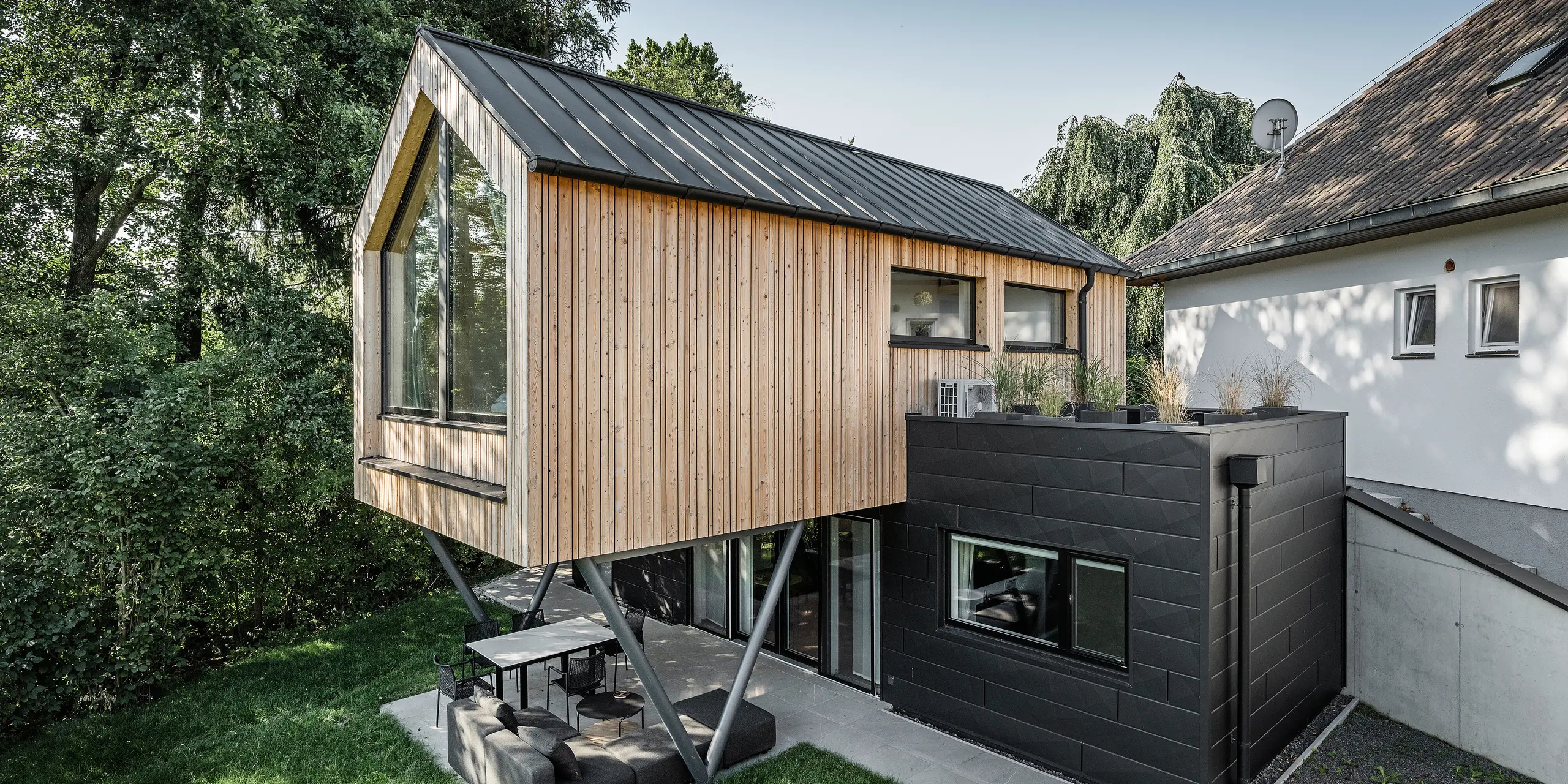 Das 'Woodstock Chalet 5' beeindruckt mit einer eindrucksvollen Kombination aus natürlichen Holzoberflächen und den modernen PREFA Siding.X Fassadenelementen in Anthrazit. Die überhängende Holzstruktur des Obergeschosses ragt markant über die dunklen Fassadenelemente, die das Erdgeschoss verkleiden, und erzeugt so ein Spiel aus Licht und Schatten. Die klaren Linien und die robuste Konstruktion der Siding.X Elemente ergänzen das innovative Design des Gebäudes und bieten einen spannenden visuellen Kontrast zur umgebenden Vegetation und dem traditionellen Gebäude im Hintergrund. Am Dach des Chalets wurde das PREFALZ Dachsystem in Form einer gleichmäßigen Stehfalzdeckung verlegt. Am rechten Rand des Erdgeschosses ist das PREFA Dachentwässerungssystem zu erkennen, das analog zur Aluminiumfassade in Anthrazit gehalten ist. Für die Dachentwässerung wurden der Wasserfangkasten gefolgt vom Ablaufrohr montiert.