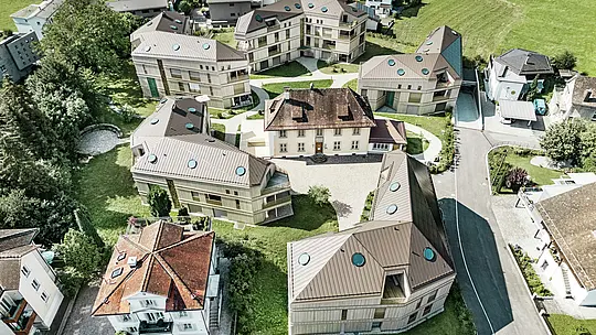 Vista aerea del moderno complesso residenziale di Liebwylen, in Svizzera.  Gli edifici sono progettati con tetti in alluminio PREFALZ di colore marrone e sono dotati di lucernari rotondi. Al centro si trova la villa storica, circondata da nuovi edifici disposti in modo armonioso e inseriti in un paesaggio verde.