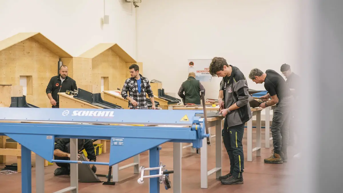 Praktische Schulung in der PREFA Academy Italien: Mehrere Teilnehmer arbeiten an Werkbänken mit Aluminiumbauteilen und -werkzeugen. Im Vordergrund ist eine blaue Biegemaschine der Marke Schechtl zu sehen.