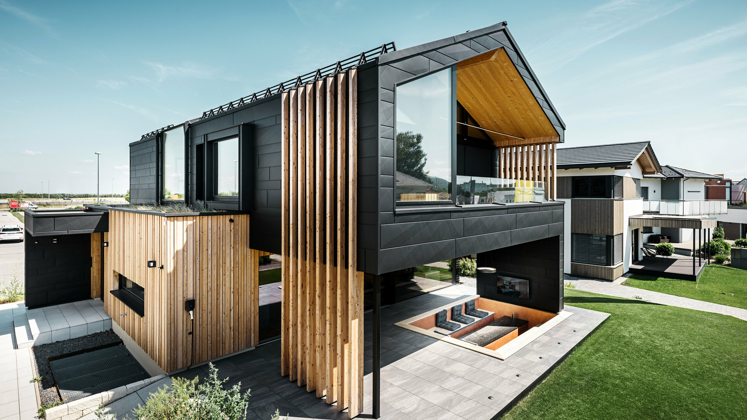 Seitenansicht des Musterhauses Autark in der Blauen Lagune in Vösendorf, Österreich. Das Obergeschoss besticht durch große Fensterflächen und eine moderne Aluminiumfassade aus PREFA Siding.X Paneelen in P.10 Schwarz, kombiniert mit vertikalen Holzelementen. Die großzügige Terrasse im Erdgeschoss mit eingelassener Sitzgelegenheit fügt sich nahtlos in die Architektur ein. Das stilvolle und nachhaltige Design des Hauses verbindet Aluminium und Holz auf eindrucksvolle Art und Weise.