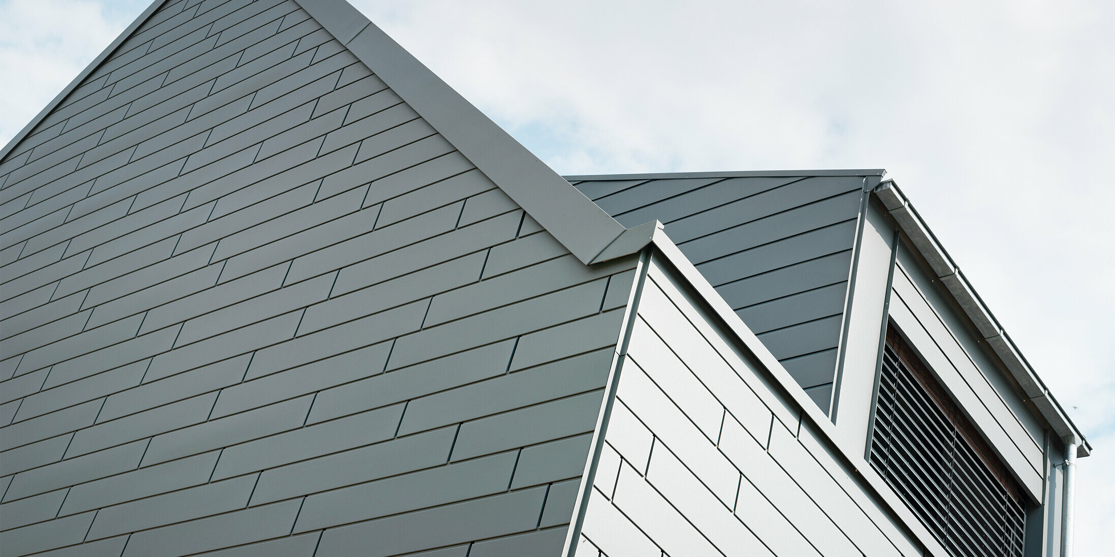 Ein Blick auf die Dachspitze des Gebäudes verrät die präzise Verarbeitung der PREFA Sidings in der Farbe P.10 Hellgrau. Das harmonische Zusammenspiel der schrägen Linien sowie das durchdachte Design der Ecken schaffen eine moderne, elegante Silhouette. Die monochrome Fassade unterstreicht die Zeitlosigkeit des Hauses.