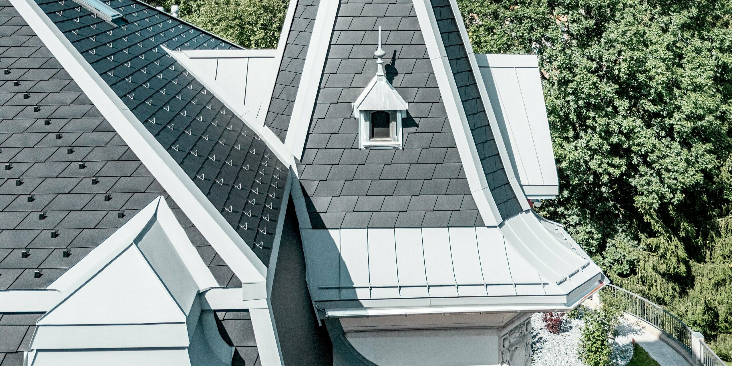 Detailaufnahme des filigranen Turms der Manufacture Horlogère du Locle in der Schweiz, der als eleganter Eyecatcher über der komplexen Dachlandschaft thront. Die steilen, präzise gedeckten Flächen aus PREFA Dachschindeln in P.10 Schwarz erzeugen eine markante Struktur, während PREFALZ in Patinagrau geschwungene Übergänge und feine Rundungen perfekt formt. Das leichte Aluminiumdach überzeugt durch hohe Witterungsbeständigkeit, außergewöhnliche Langlebigkeit und maximale Gestaltungsfreiheit für anspruchsvolle, architektonisch einzigartige Dachlösungen.