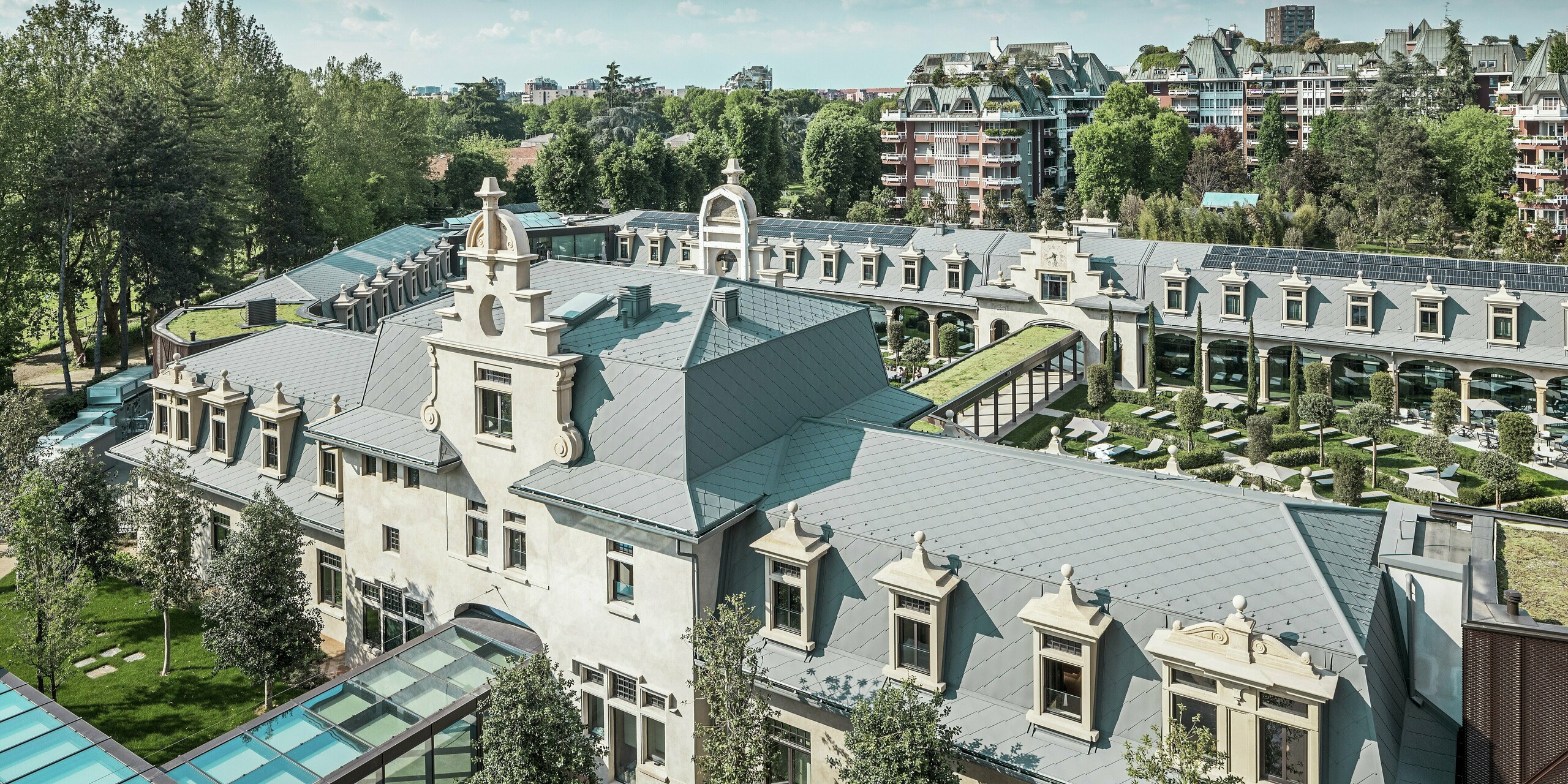 Gesamtansicht der imposanten Anlage „De Montel – Terme Milano“ mit denkmalgeschützter Jugendstilarchitektur und modernem Wellnessambiente. Die großflächige Dacheindeckung mit PREFA Dachrauten 44 × 44 in P.10 Hellgrau verleiht dem Komplex eine elegante Einheitlichkeit. Integrierte PV-Module, begrünte Dächer und Glasverbindungen zwischen den Baukörpern spiegeln den Anspruch an Nachhaltigkeit und zeitgemäßes Design im urbanen Kontext wider.
