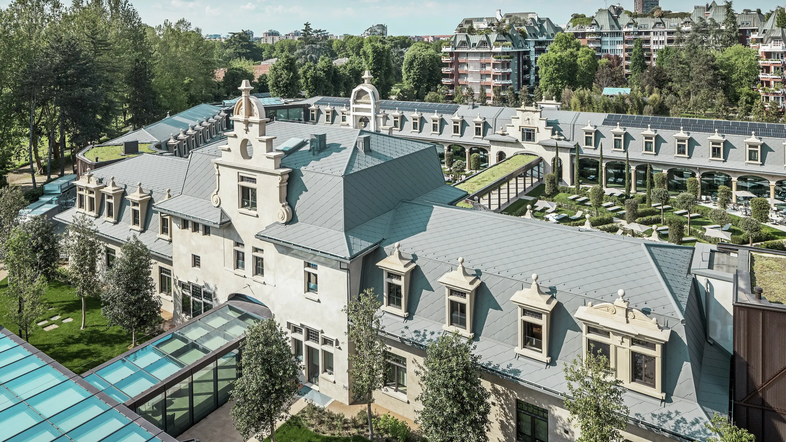 Vista panoramica dell'imponente complesso “De Montel – Terme Milano” con architettura Art Nouveau protetta come monumento storico e moderno ambiente benessere. L'ampia copertura del tetto con scaglie PREFA 44 × 44 in P.10 grigio chiaro conferisce al complesso un'elegante uniformità. I moduli fotovoltaici integrati, i tetti verdi e i collegamenti in vetro tra gli edifici riflettono l'esigenza di sostenibilità e design contemporaneo nel contesto urbano.