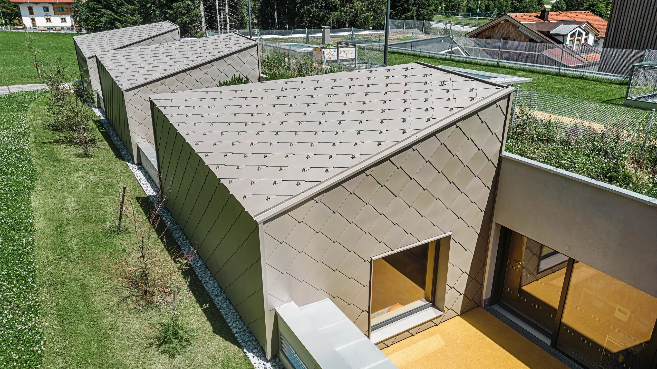 Le scaglie PREFA 44 × 44, color bronzo, rivestono il tetto e parte della facciata di un edificio moderno a Gerlos, nella Zillertal. L'edificio è dotato di ampie vetrate e terrazze. Sullo sfondo si intravede l'ambiente rurale con prati verdi e una collina boscosa.