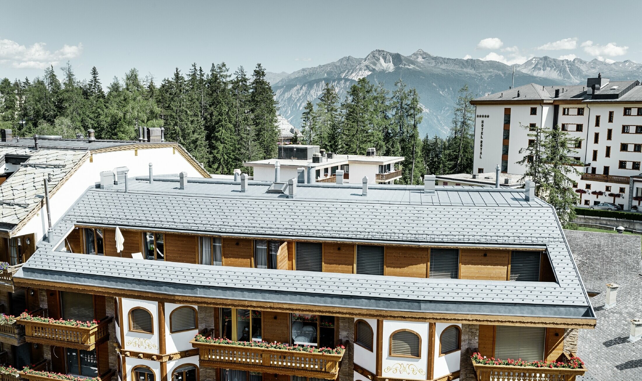 Casa plurifamiliare in Crans-Montana con montagne sullo sfondo e una facciata con elementi decorativi in legno e con una copertura in scandole PREFA grigio pietra