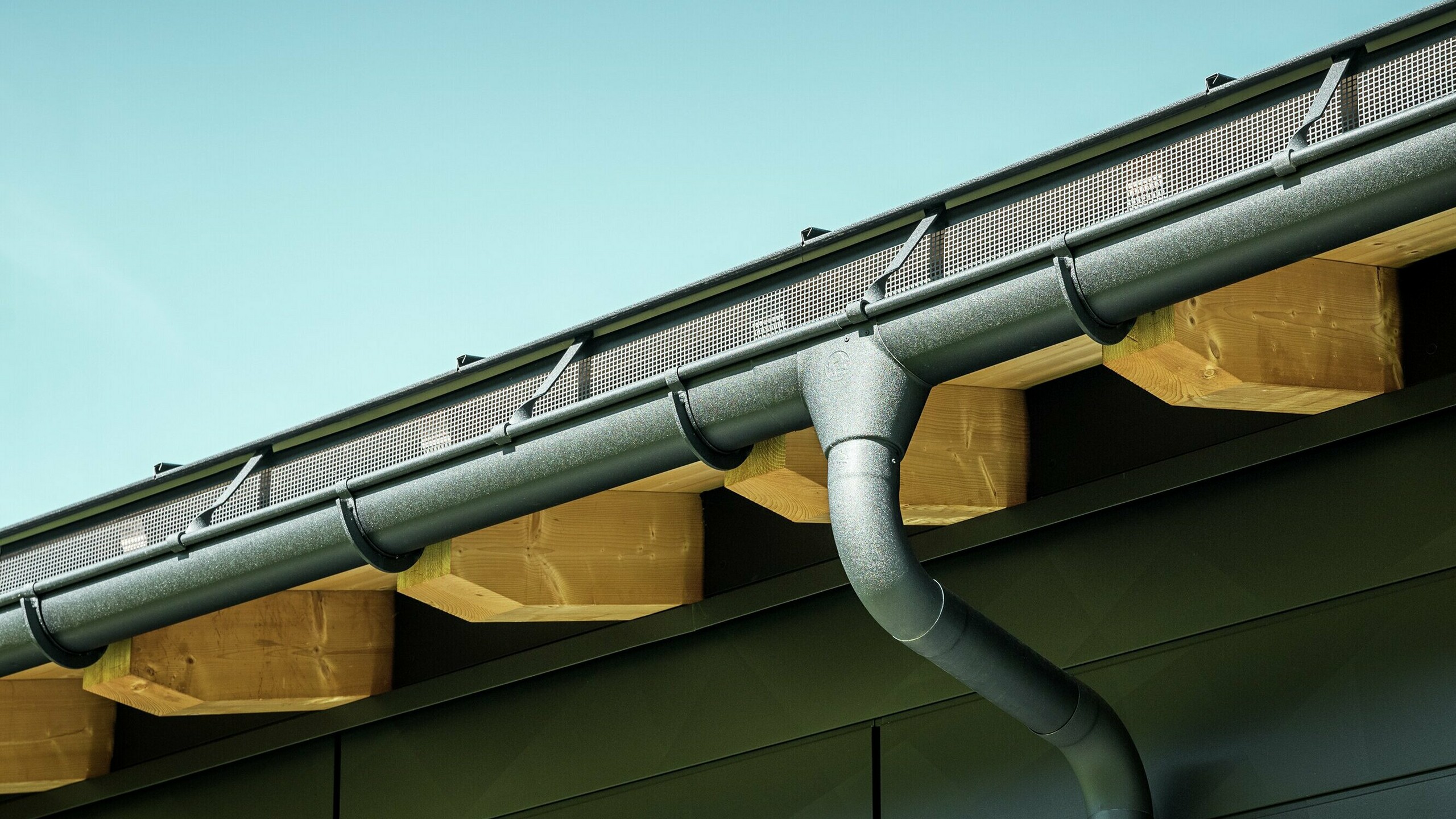 PREFA Hängerinne mit Rinnenkessel und Überbügel in P.10 Schwarz Nahaufnahme der Fassade eines modernen Einfamilienhauses in Ascharina, Schweiz. Die Gebäudeseiten wurden mit den markanten PREFA Siding.X Fassadenpaneelen in P.10 Schwarz verkleidet. Farblich abgestimmt ist die PREFA Dachentwässerung mit Hängerinne und Ablaufrohr.