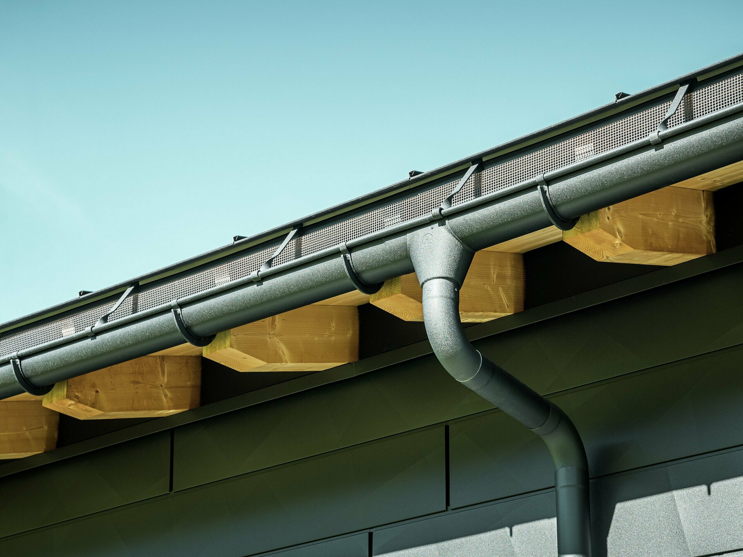 Nahaufnahme der Fassade eines modernen Einfamilienhauses in Ascharina, Schweiz. Die Gebäudeseiten wurden mit den markanten PREFA Siding.X Fassadenpaneelen in P.10 Schwarz verkleidet. Farblich abgestimmt ist die PREFA Dachentwässerung mit Hängerinne und Ablaufrohr.