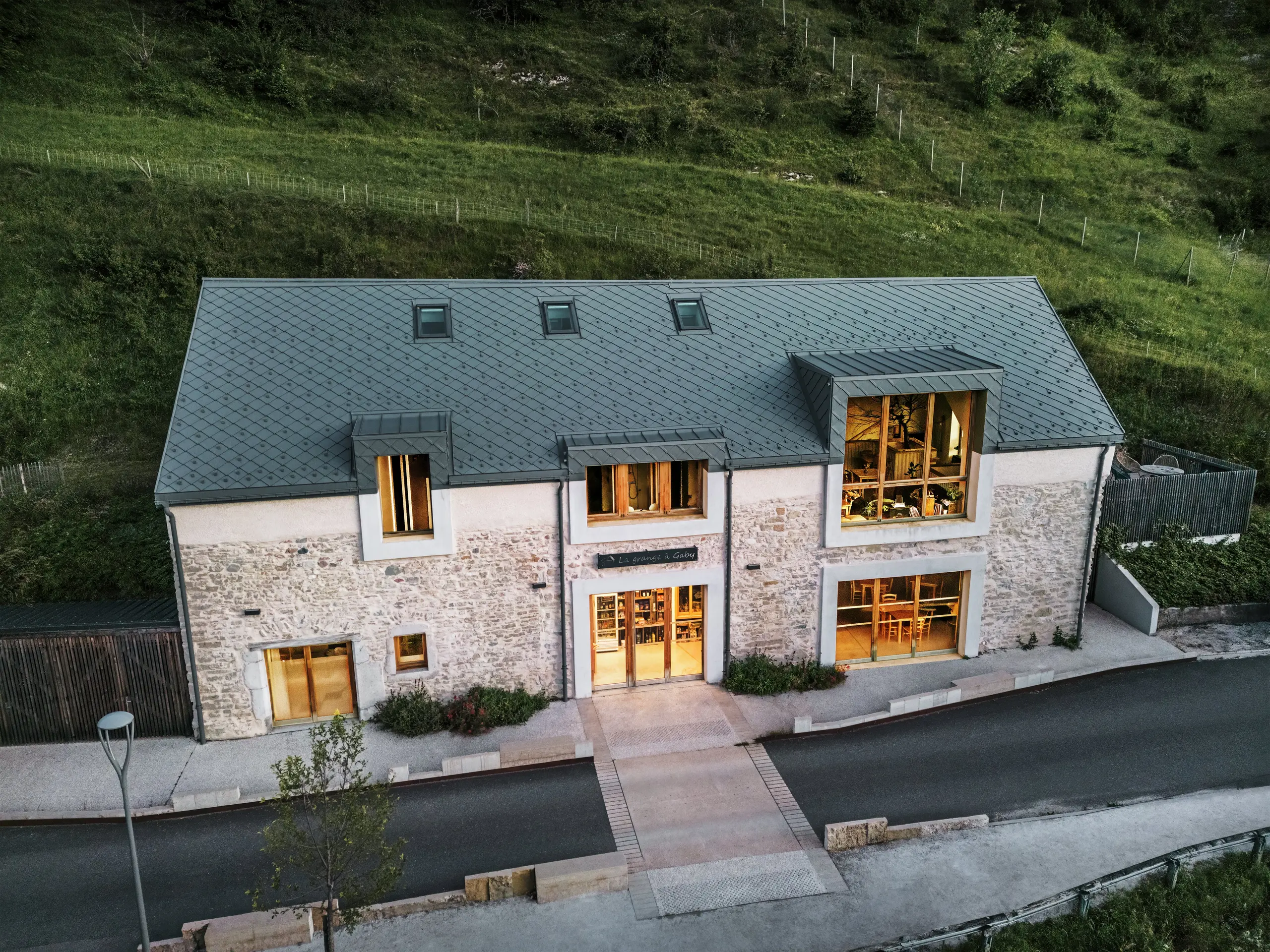 Abendansicht der renovierten Scheune „La Grange à Gaby“ in Engins, Frankreich. Das Gebäude vereint traditionelles Steinmauerwerk mit einem modernen PREFA-Aluminiumdach mit 29 × 29 cm großen Rauten. Große Fenster und helle Innenräume unterstreichen die behagliche Atmosphäre des neuen Kulturzentrums.