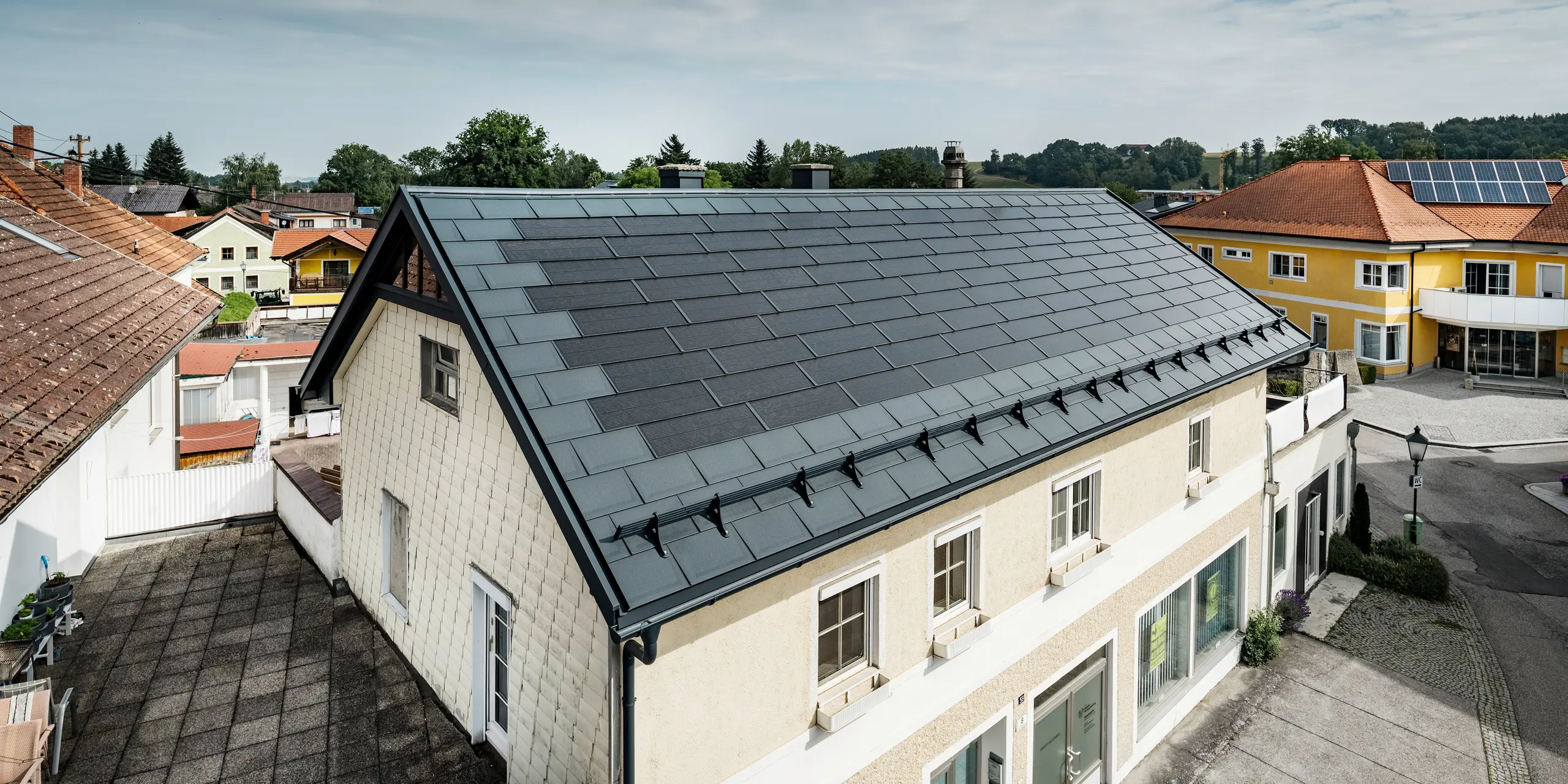 Vista dall'alto di una casa indipendente a Mettmach, in Austria. Il vecchio edificio è stato ristrutturato con tegole fotovoltaiche PREFA color antracite. I sistemi di protezione dalla neve e le linee chiare delle tegole fotovoltaiche sottolineano la lavorazione pulita dei prodotti in alluminio PREFA. Il tetto a tegole di colore rosso di un edificio vicino contrasta sullo sfondo. I moduli fotovoltaici sono installati sul tetto della vicina casa unifamiliare in una classica soluzione su tetto, a dimostrazione della diversità della progettazione di tetti ad alta efficienza energetica.