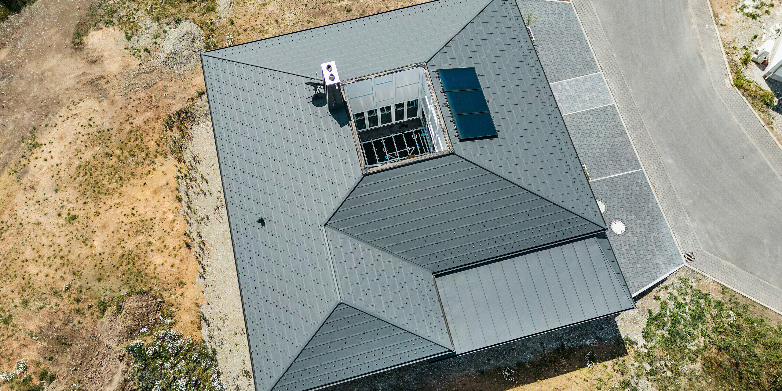 Luftbild eines modernen Einfamilienhauses in Ilmenau, Thüringen, mit einem Aluminiumdach aus PREFA Dachplatten R.16 in P.10 Dunkelgrau. Die präzise verlegten Dachplatten und das nahtlos anschließende PREFALZ Dachsystem über der Garage erzeugen ein harmonisches Gesamtbild. Das Dach des Bungalows hebt sich mit seiner klaren Linienführung und dem zentralen Atrium, umrahmt von der Qualität und Ästhetik der PREFA Aluminiumprodukte, hervor.