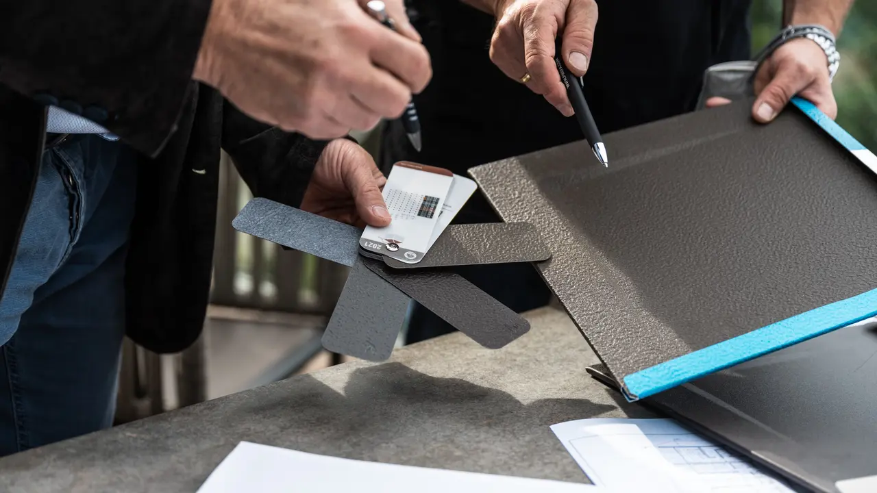 Zwei Personen vergleichen einen Farbfächer mit mehreren Grautönen und zeigen auf eine PREFA Dachraute aus Aluminium, die auf einem Tisch liegt; daneben liegen Planungsunterlagen