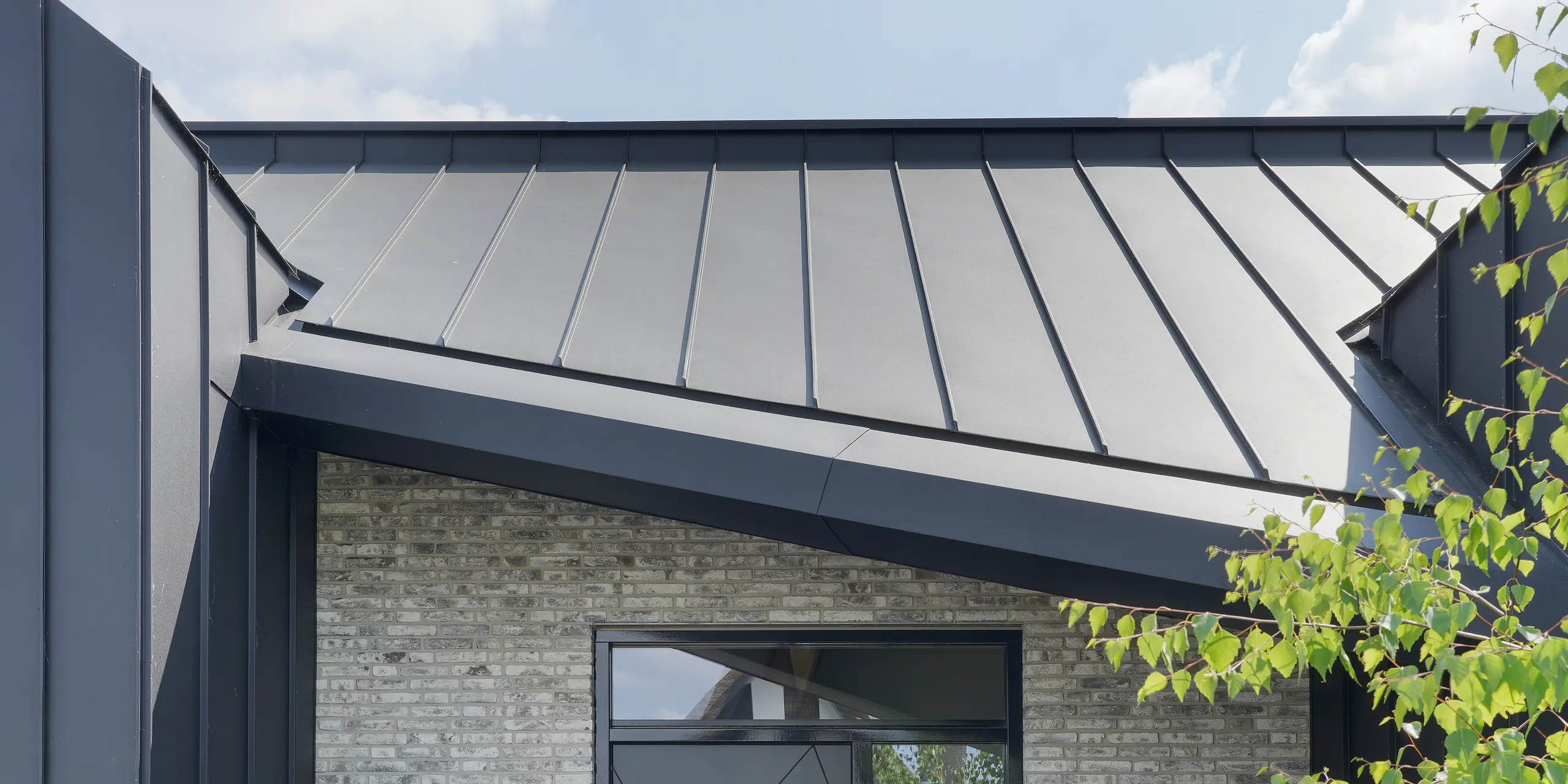 Eingangsbereich eines stilvollen Familienhauses in Veenendaal, Niederlande. Der Eingang wird durch eine schwarze Aluminiumverkleidung hervorgehoben. Das PREFALZ Dach- und Fassadensystem kontrastiert mit dem hellgrauen Naturstein der Fassade. Die große schwarze Tür trägt die Hausnummer 40 und bietet einen eleganten Willkommenspunkt neben der attraktiv gestalteten Gartenlandschaft.