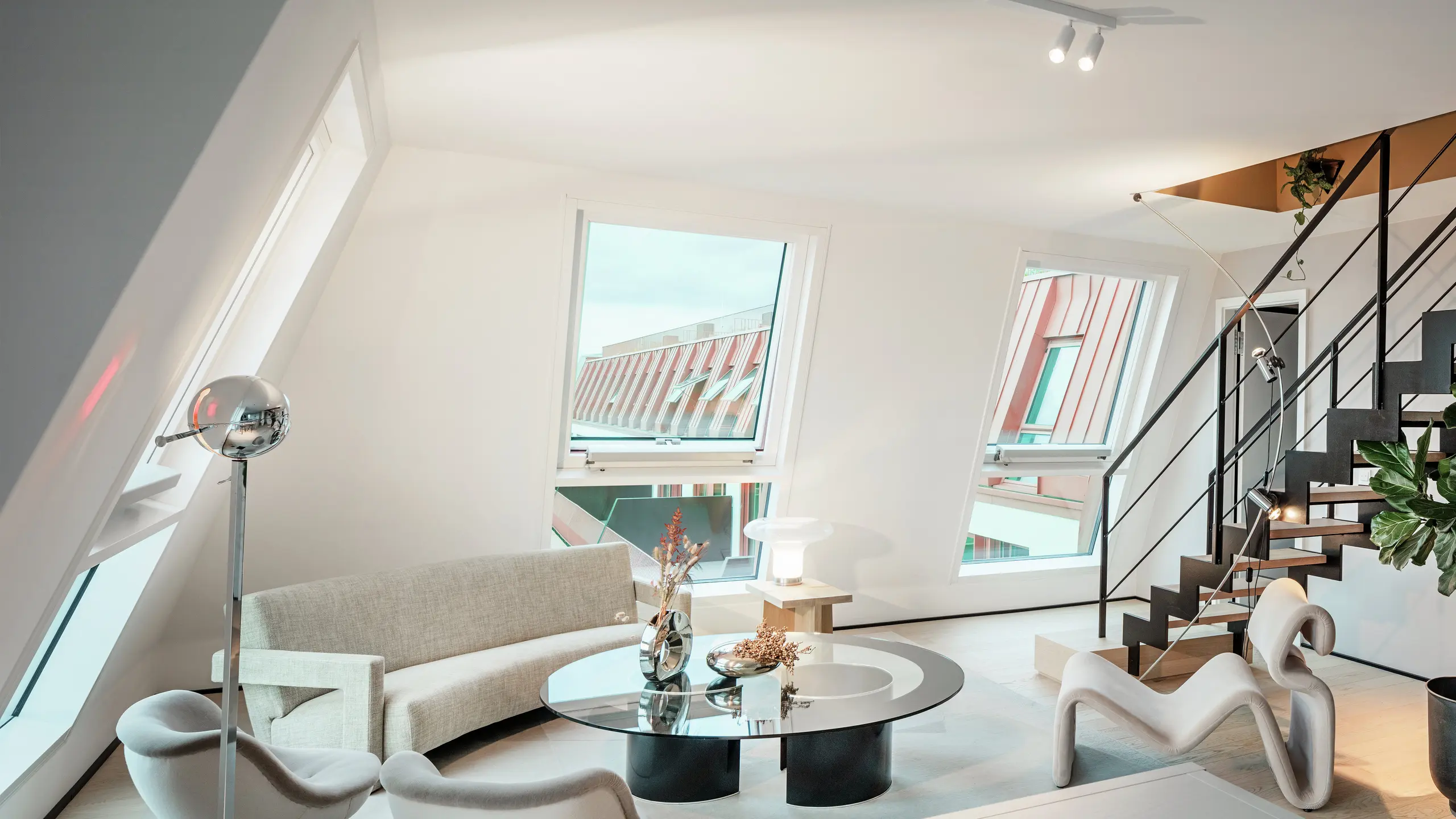 Vista interna di un attico inondato di luce nel progetto residenziale Van B a Monaco di Baviera, con una vista del tetto in alluminio PREFALZ di alta qualità nel colore speciale IGP Orange HDP.  Le ampie finestre lasciano entrare molta luce naturale e sottolineano il moderno concetto abitativo. Le linee chiare, i mobili di design e la precisa lavorazione dei collegamenti del tetto dimostrano come l'architettura di alta qualità e di stile possa essere realizzata non solo all'esterno, ma anche all'interno.