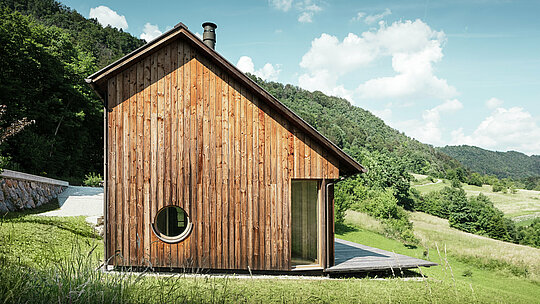 Casa minimalista in legno con apertura a finestra rotonda e tetto in alluminio PREFALZ di colore marrone P.10, immersa nel paesaggio collinare di Vinje. La copertura del tetto PREFA non richiede manutenzione, offre una protezione ottimale contro il vento e le intemperie e si armonizza nel colore con la facciata in legno naturale. Sottolinea l'architettura chiara e senza tempo dell'edificio. Dalla spaziosa terrazza si gode di una meravigliosa vista sulle colline slovene. La prospettiva mostra la facciata della casa indipendente con il suo tetto a falde asimmetriche e le superfici del tetto di diverse dimensioni.