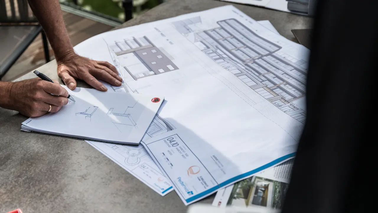 Eine Person skizziert Entwürfe auf einem Notizblock, während große Baupläne auf einem Tisch ausgebreitet liegen; die Pläne zeigen architektonische Details eines Gebäudes.