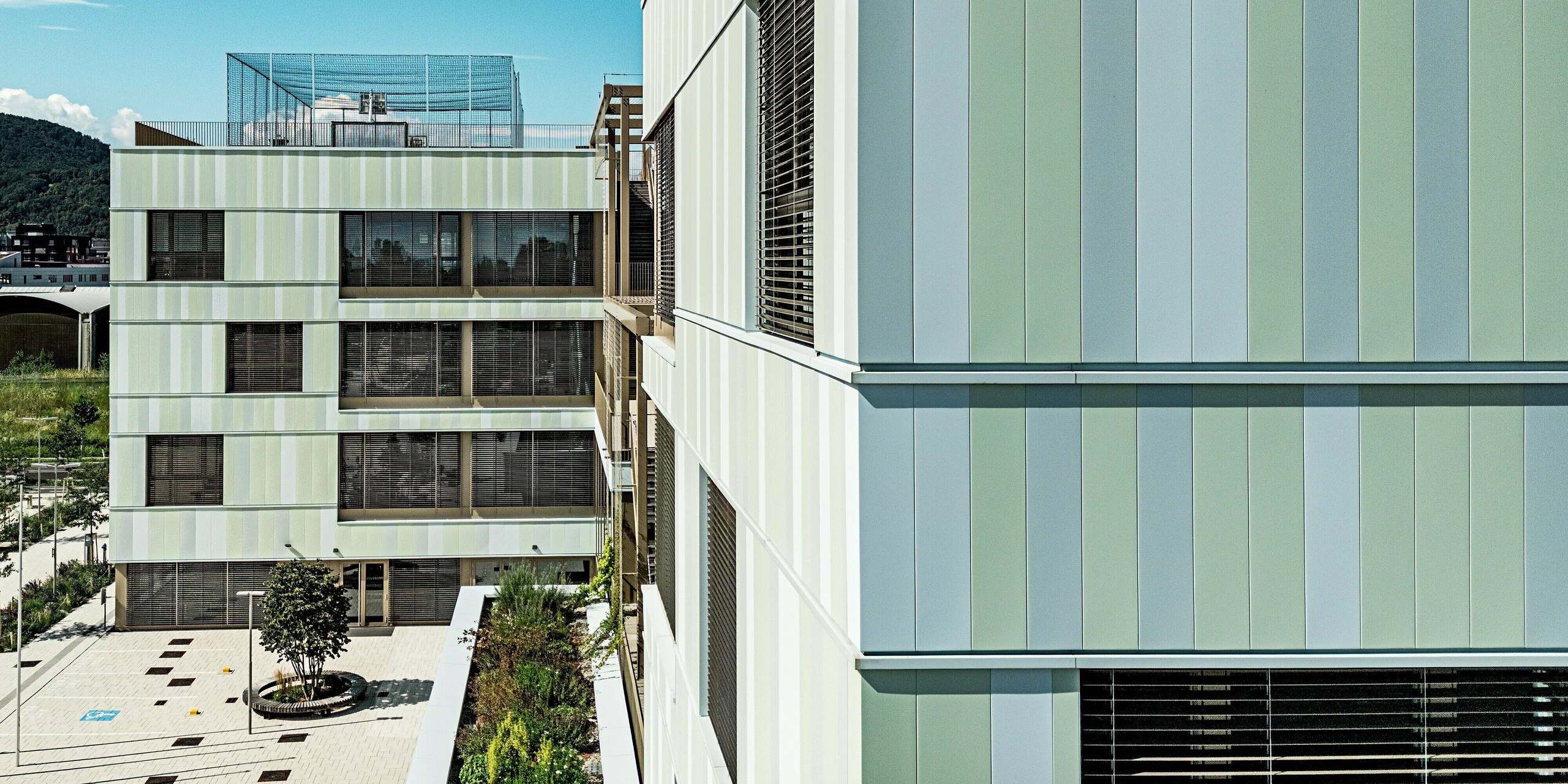 Das Bild zeigt die moderne Fassade der AHS Reininghaus in Graz, die durch ihre auffälligen PREFA-Sidings in Sonderfarben besticht. Die in zarten Grüntönen und Weiß gehaltenen Fassadenelemente verleihen dem Gebäude eine elegante, zugleich aber auch dynamische Ausstrahlung. Die Fenster sind mit dunklen Jalousien ausgestattet, die nicht nur einen funktionalen Sonnenschutz bieten, sondern auch einen ästhetischen Kontrast zur helleren Fassade bilden. Das Gebäude fügt sich perfekt in die umgebende Landschaft ein. Die Kombination aus modernen Materialien und durchdachtem Design macht die Schule zu einem Blickfang in der Grazer Stadtlandschaft.