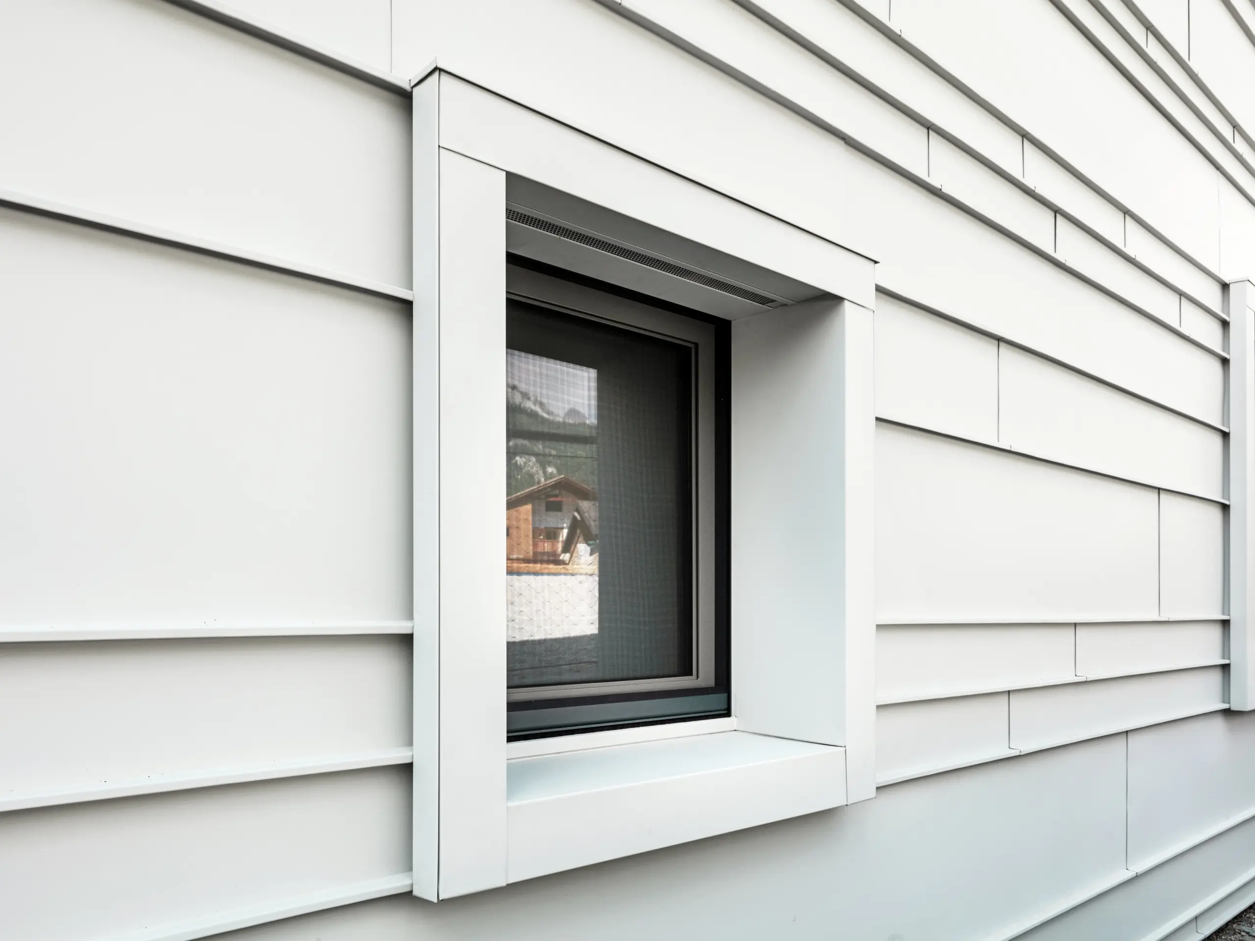 Dettaglio di una finestra caratteristica in una facciata ventilata realizzata con PREFALZ P.10 nel colore bianco PREFA. Le lamelle in alluminio posate in orizzontale conferiscono struttura, chiarezza e un'estetica moderna. I telai delle finestre della caserma “Tempesti”, che spiccano in modo particolare, iniziano e terminano sempre esattamente in corrispondenza di una linea di aggraffatura. In questo modo viene volutamente enfatizzata la struttura dell'alluminio.