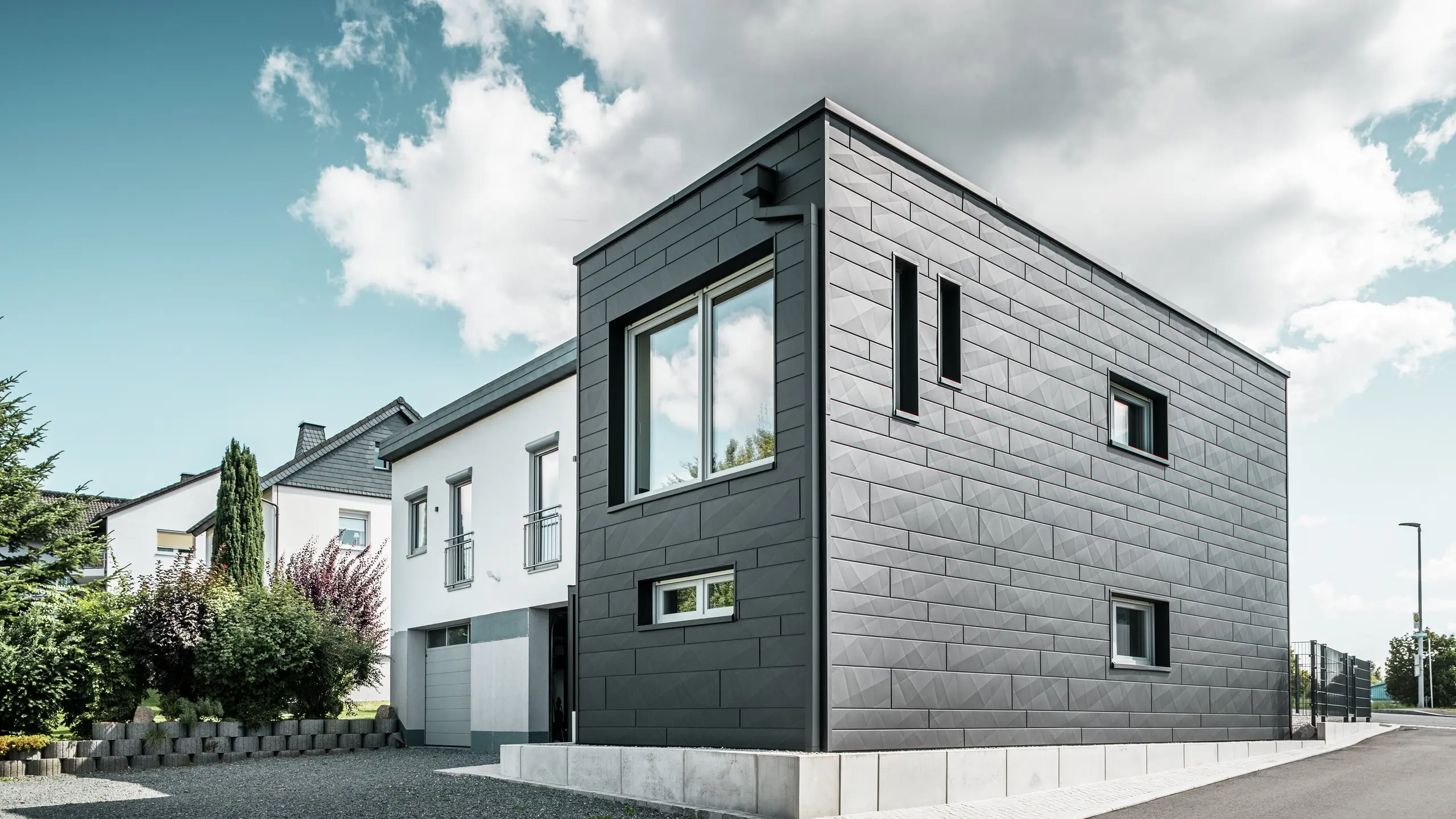 La facciata elegante e moderna della casa unifamiliare ristrutturata a Teuschnitz presenta i pannelli PREFA Siding.X in P.10 nero dalla struttura chiara. La disposizione geometrica dei pannelli in lamiera di alta qualità conferisce all'edificio un'estetica distintiva e senza tempo. Le finestre sono incorniciate da pannelli compositi in alluminio nero, che completano armoniosamente il design. Il sistema di smaltimento acque piovane PREFA integrato con pluviale quadro offre una soluzione funzionale ed esteticamente gradevole.
