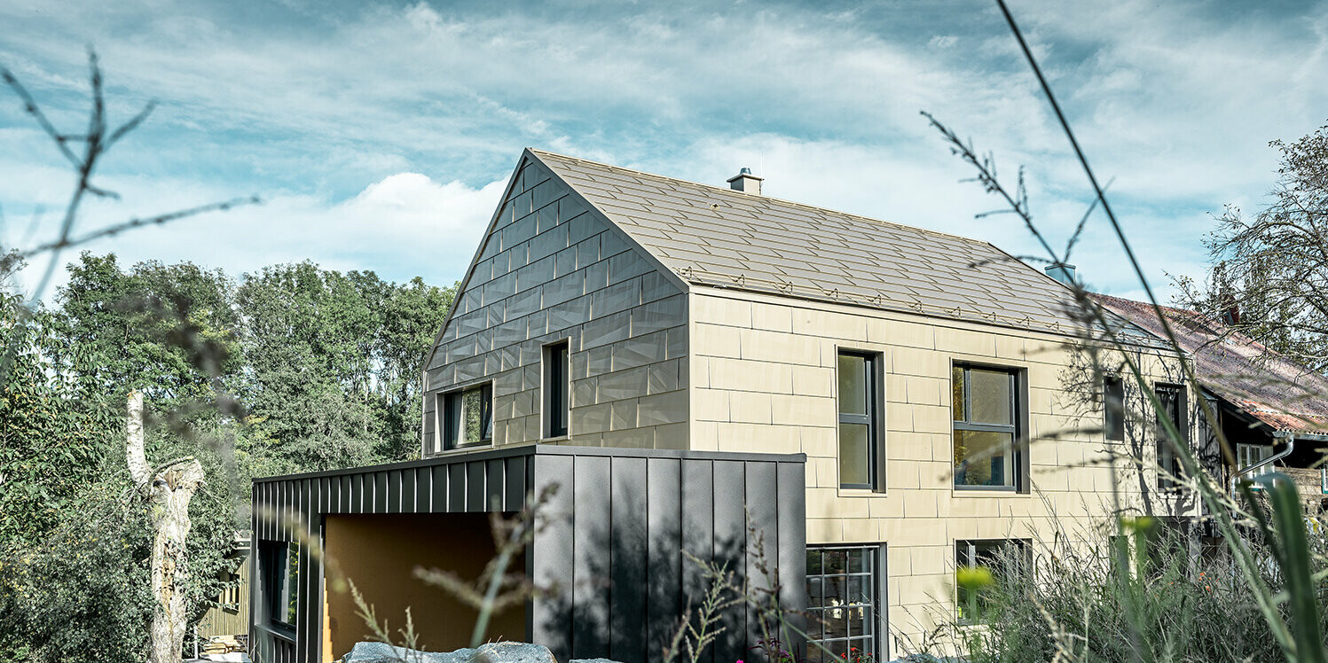 Wohnhaus ohne Dachvorsprung ist am Dach und der Fassade mit dem Aluminium Paneel FX.12 von PREFA in Bronze verkleidet. Gleich daneben steht eine Garage mit Flachdach mit einer Winkelstehfalzfassade.