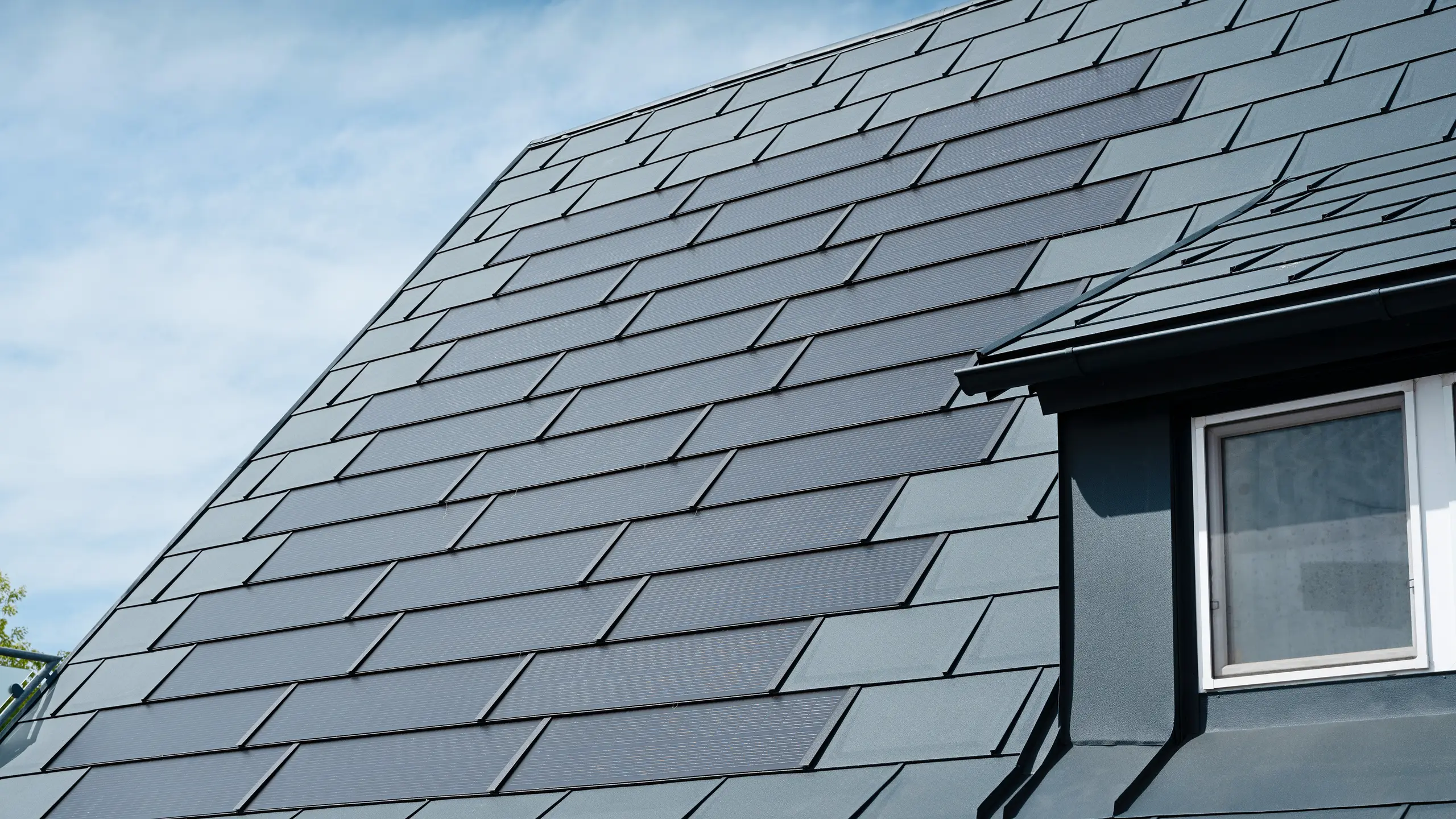 Detailansicht des sanierten Dachs eines Einfamilienhauses in Leonding mit PREFA Solardachplatten in Anthrazit. Die Nahaufnahme zeigt die hochwertigen Aluminiumplatten von PREFA, die eine nahtlose Integration der Solarmodule ermöglichen. Diese langlebige und witterungsbeständige Lösung bietet eine nachhaltige und energieeffiziente Möglichkeit zur Stromerzeugung. Das Bild verdeutlicht die ästhetische und funktionale Qualität der verwendeten Materialien. Am rechten Bildrand ist ein weißes Fenster unter einer Schleppgaube zu erkennen.