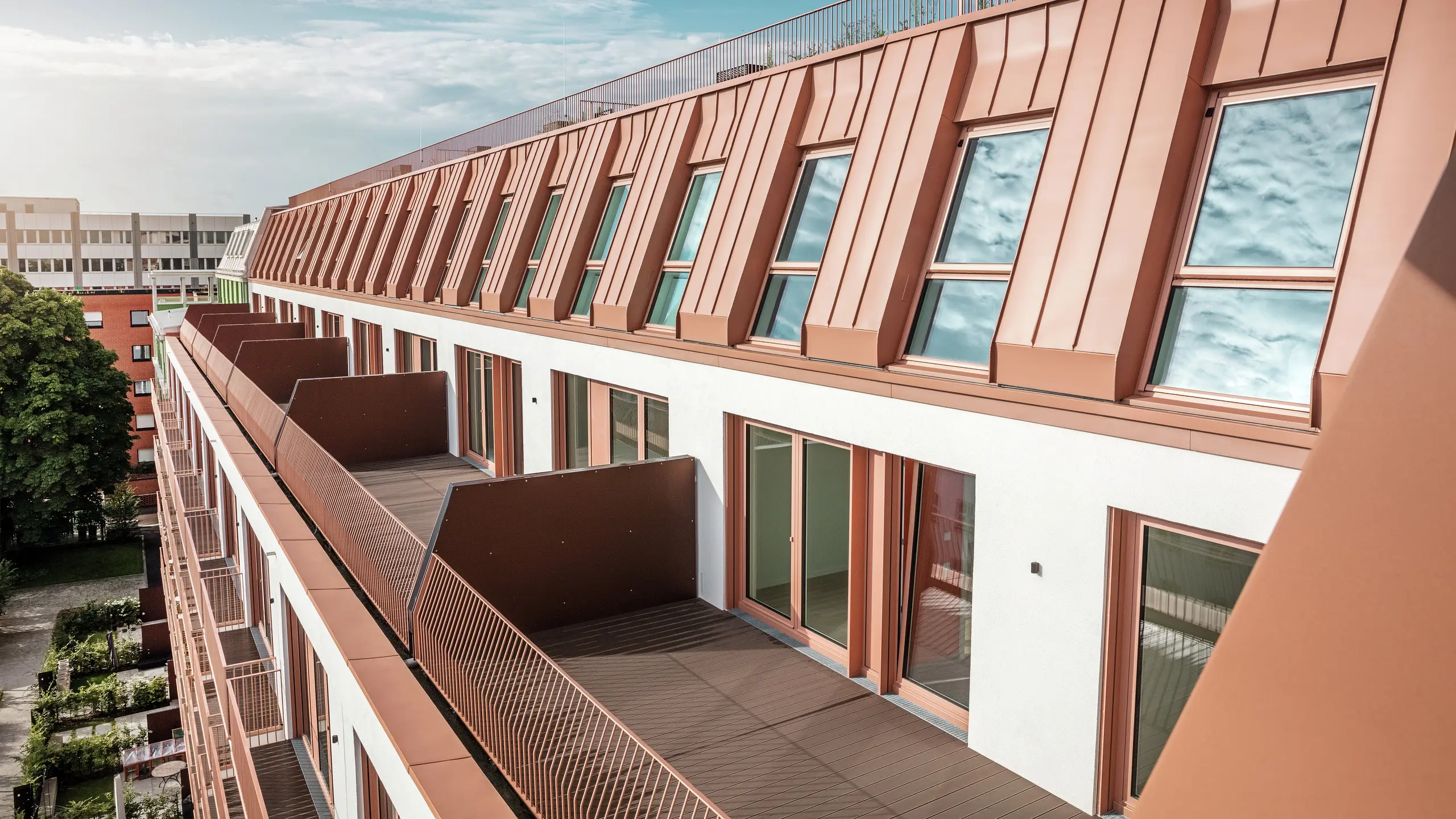 Vista dettagliata del moderno design del tetto e della facciata del progetto residenziale Van B a Monaco di Baviera con il tetto in alluminio PREFALZ di alta qualità in IGP Orange HDP di PREFA.  L'accurata lavorazione del materiale, durevole e a bassa manutenzione, offre una protezione ottimale dalle intemperie e conferisce all'edificio un aspetto uniforme ed elegante. Le ampie finestre e i balconi forniscono molta luce naturale e creano un ambiente abitativo aperto e vivibile con raffinatezza architettonica.