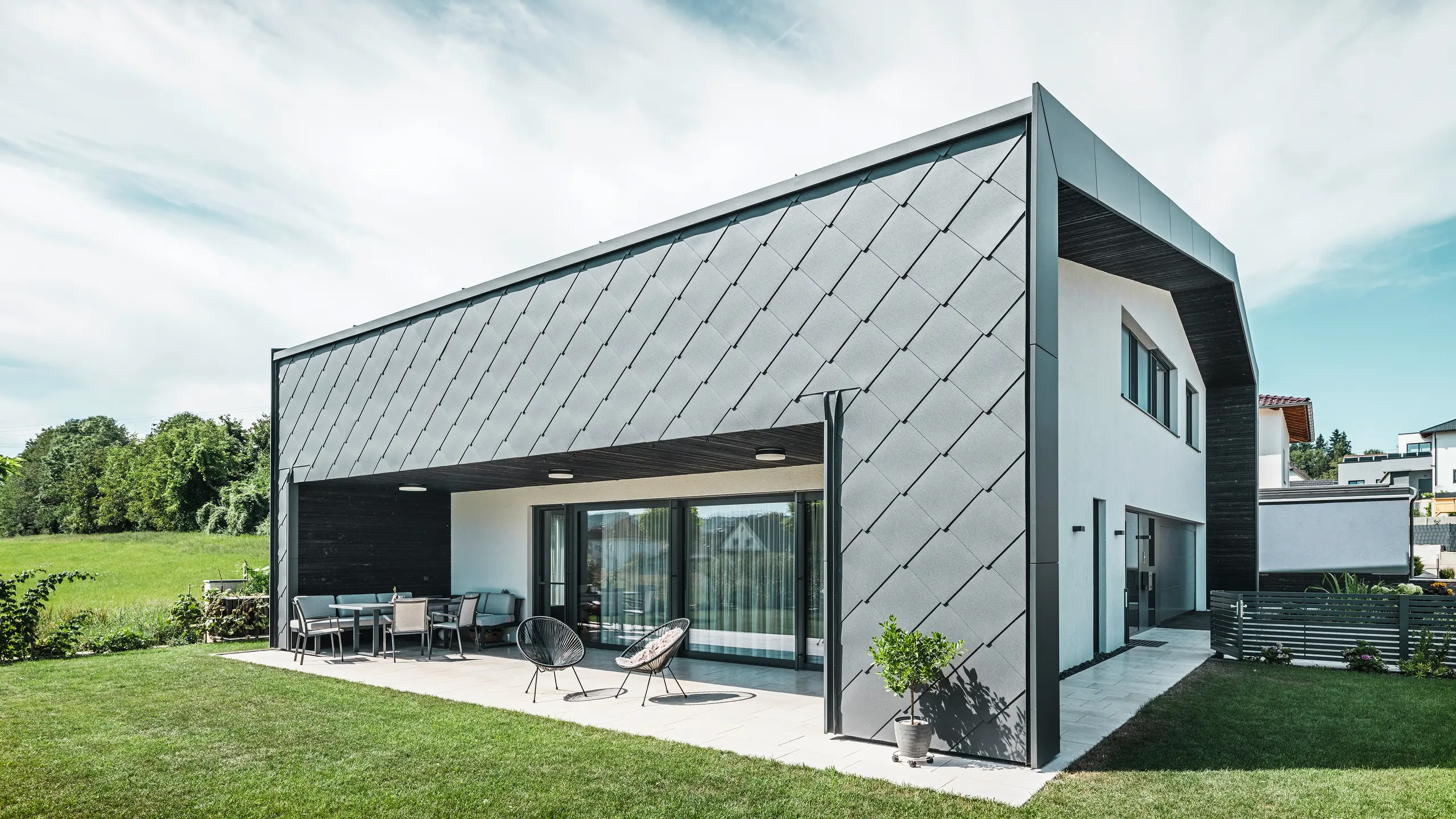 Gepflegter Gartenbereich eines Einfamilienhauses in Aschach. Das Gebäude ist stilvoll eingedeckt mit dunkelgrauen PREFA Aluminium-Wandrauten 44 × 44. Die überdachte Terrasse bietet einen gemütlichen Sitzplatz mit Blick auf die saftig grüne Rasenfläche. Die elegante Aluminiumverkleidung in Kombination mit weißen Putzflächen sorgt für eine moderne und witterungsbeständige Architektur. Große Fensterflächen lassen viel Tageslicht in den Innenraum und schaffen eine offene, lichtdurchflutete Atmosphäre.