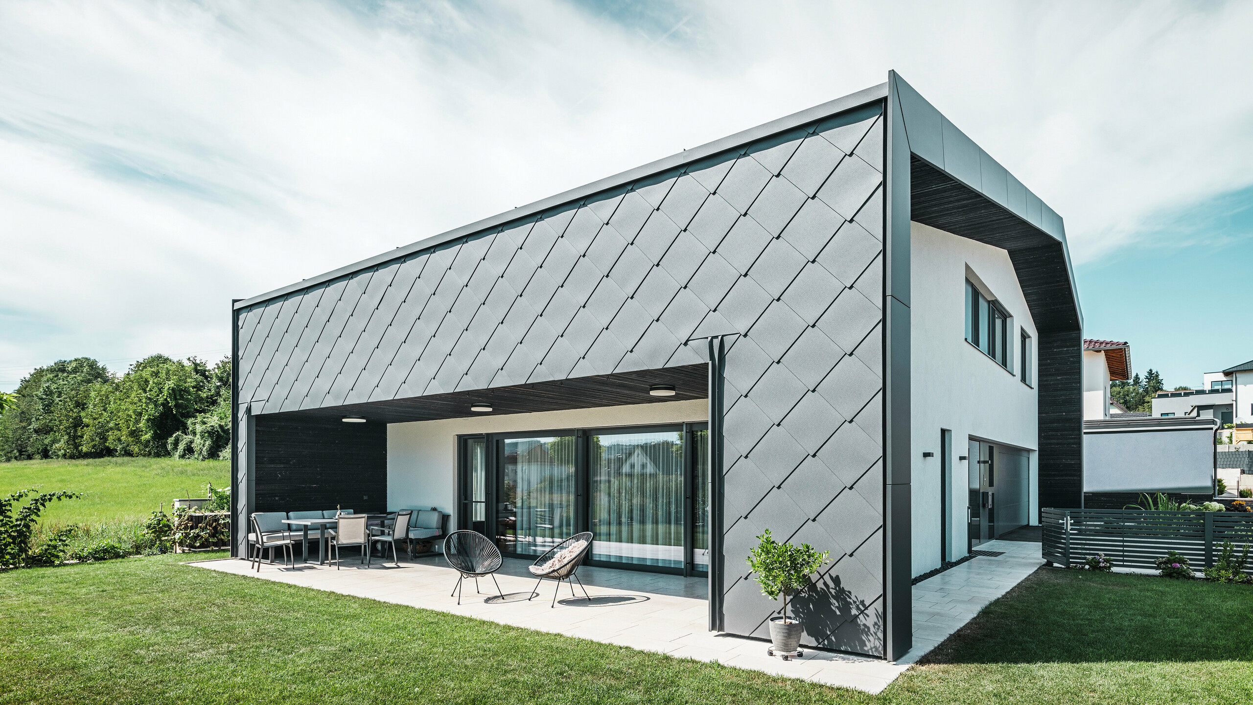 Giardino ben curato di una casa unifamiliare ad Aschach. L'edificio è elegantemente rivestito con scaglie per facciata PREFA in alluminio grigio scuro 44 × 44. La terrazza coperta offre un accogliente posto a sedere con vista sul prato verde e rigoglioso. L'elegante rivestimento in alluminio, in combinazione con le superfici intonacate bianche, garantisce un'architettura moderna e resistente alle intemperie. Le grandi superfici vetrate lasciano entrare molta luce naturale all'interno e creano un'atmosfera aperta e luminosa.