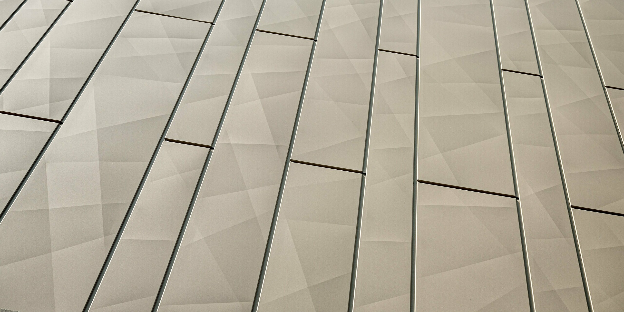 Das Bild zeigt einen Nahaufnahme der Siding-X-Paneele der Aluminiumfassade des Einkaufszentrums in Rohrbach. Die in einem eleganten Bronzeton gehaltenen Fassadenelemente zeichnen sich durch eine geometrische Struktur aus, die durch präzise Linien und flache Falten eine dynamische, dreidimensionale Textur erzeugt. Die nahtlosen Übergänge und die feine Unterteilung der Paneele vermitteln eine moderne, elegante Optik. Diese Fassadenelemente sind nicht nur ästhetisch ansprechend, sondern auch äußerst widerstandsfähig gegen äußere Einflüsse, wodurch sie besonders pflegeleicht und langlebig sind.