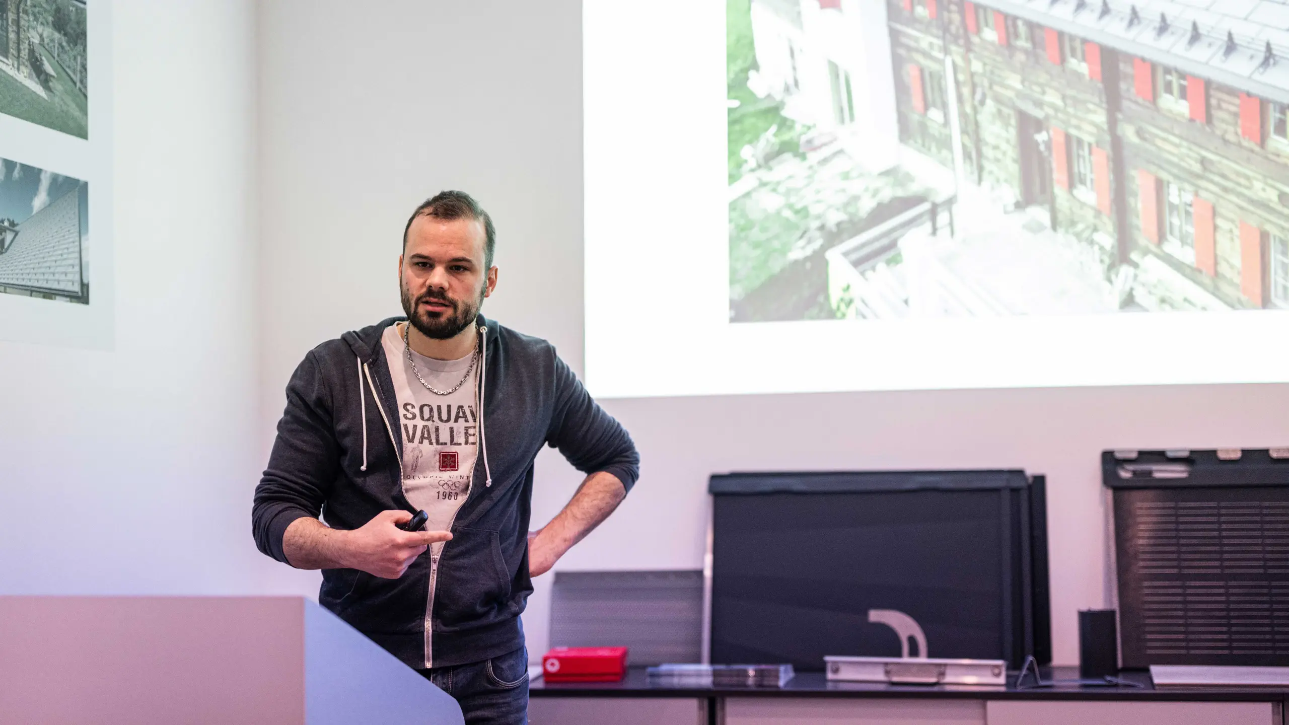 Ein PREFA Mitarbeiter hält in der PREFA Academy Italien eine Präsentation. Er steht vor einer Projektionsfläche und erklärt den Teilnehmern ein Beispielprojekt mit PREFA Dachsystemen. Im Hintergrund ist ein Bild eines Gebäudes mit PREFA Solardachplatte zu sehen. Der Vortragende spricht engagiert und vermittelt technische Details zur Anwendung der Produkte.
