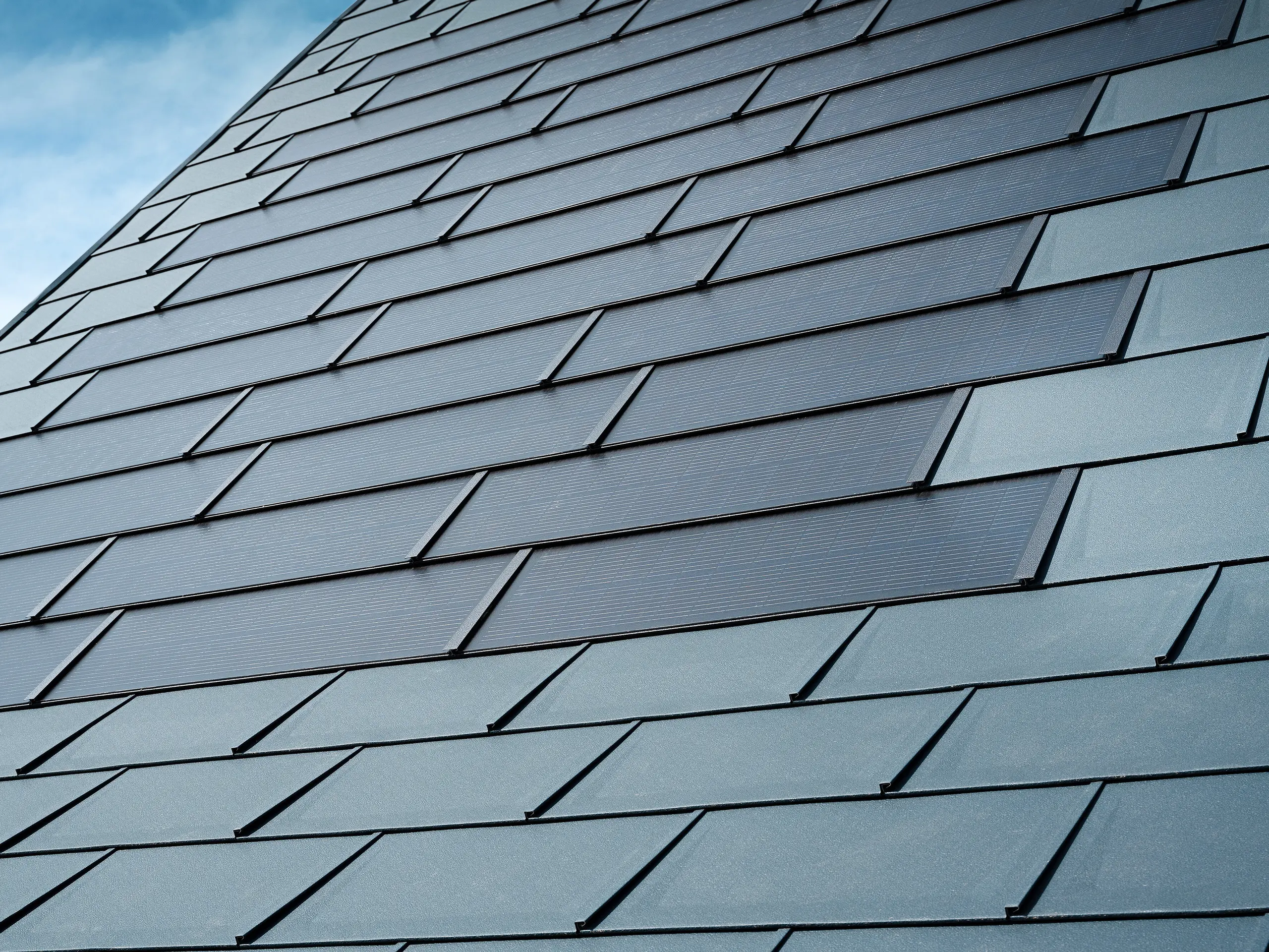 Dettaglio del tetto ristrutturato di una casa unifamiliare a Leonding con tegole fotovoltaiche PREFA in antracite. Il primo piano mostra le pregiate tegole in alluminio PREFA, che consentono un'integrazione perfetta dei moduli fotovoltaici. Questa soluzione durevole e resistente alle intemperie offre un'opzione sostenibile ed efficiente dal punto di vista energetico per la produzione di elettricità. L'immagine evidenzia la qualità estetica e funzionale delle lamiere di alluminio utilizzate. Intorno alle innovative tegole fotovoltaiche sono state posate tegole R.16 dello stesso colore.