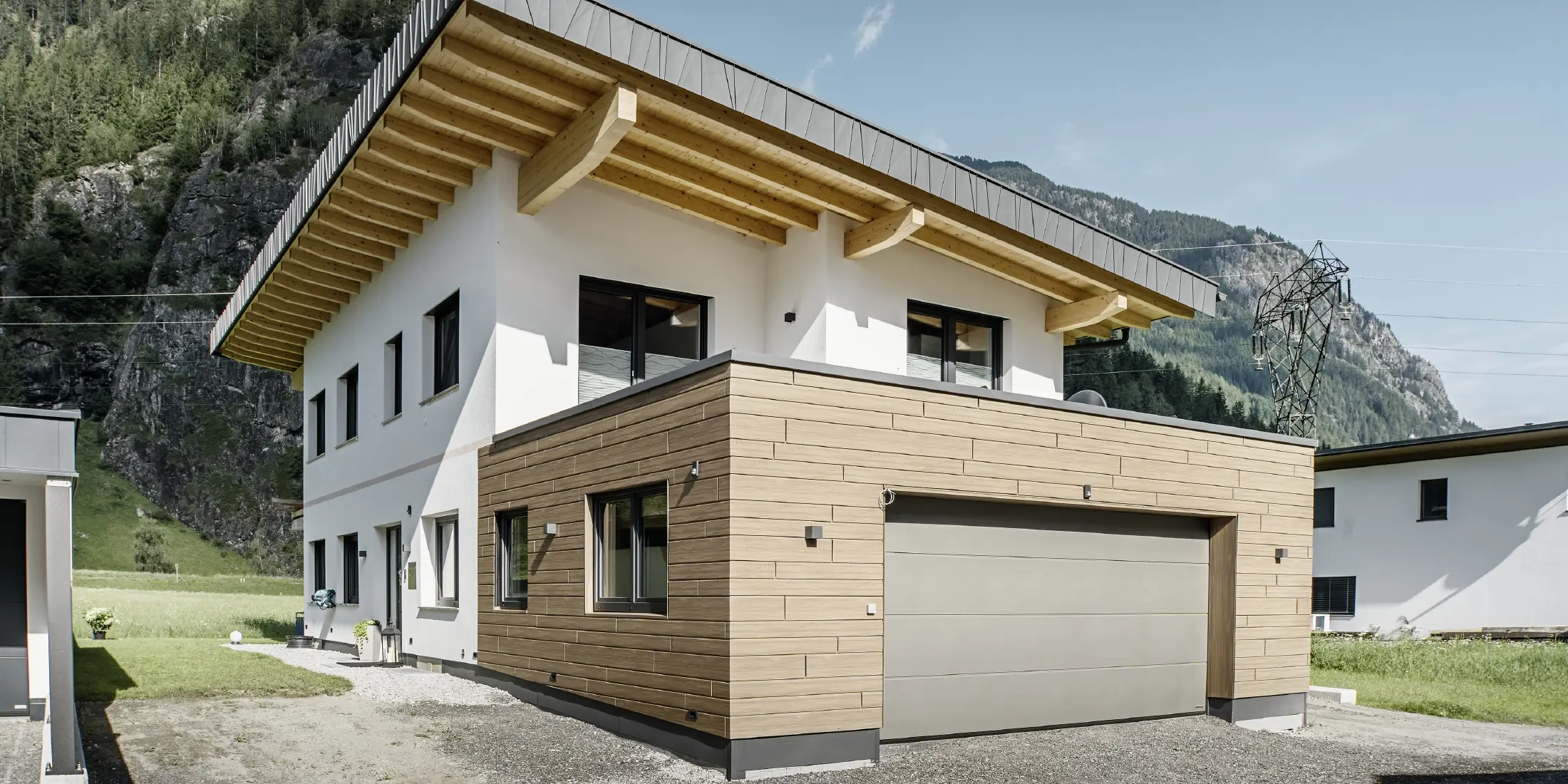 Moderna casa unifamiliare con facciata ristrutturata: facciata esterna rinnovata con rivestimento orizzontale effetto legno, abbinato a superfici intonacate di bianco, rivestimento del garage integrato e architettura contemporanea in un contesto alpino.