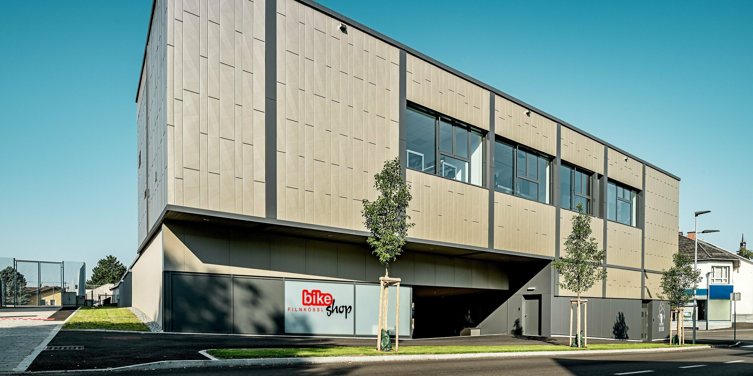 Das Bild zeigt das moderne Einkaufszentrum in Rohrbach mit seiner markanten Aluminiumfassade aus PREFA Siding X-Paneelen in Bronze. Der Gebäudekomplex beeindruckt durch seine zeitgemäße Struktur. Die markanten Fassadenelemente setzen nicht nur optische Akzente, sondern bieten auch zuverlässigen Schutz vor Witterungseinflüssen. Der Bike Shop Filnkössl und das Eiscafé Delphin bieten verschiedene Freizeitmöglichkeiten in der kleinen Gemeinde in Niederösterreich.