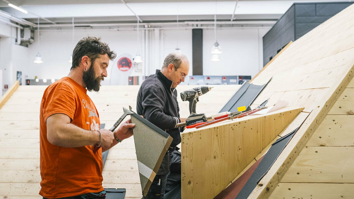 Due partecipanti alla PREFA Academy Italia lavorano su un tetto inclinato con sottostruttura in legno. L'uomo in primo piano tiene in mano un pannello per tetto PREFA tagliato su misura, mentre l'altro lavora al montaggio con un avvitatore a batteria e altri attrezzi. La scena mostra una formazione pratica sulla lavorazione a regola d'arte dei sistemi per copertura PREFA.