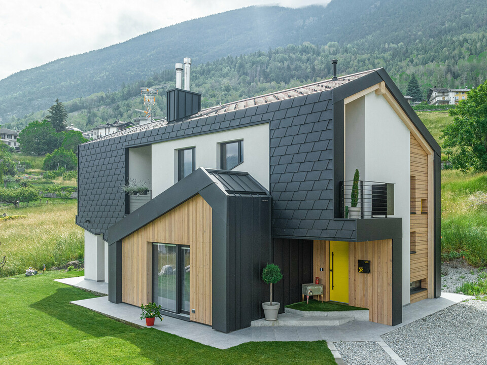 Moderne Architektur trifft auf Nachhaltigkeit in diesem Einfamilienhaus, das mit PREFA Dachschindeln DS.19 und Wandschindeln in P.10 Schwarz ausgestattet ist. Die Schindeln bilden einen starken Kontrast zu den hellen Holzelementen und der umgebenden grünen Landschaft, während das Haus selbst mit seiner klaren Linienführung und dem ansprechenden Design hervorsticht.