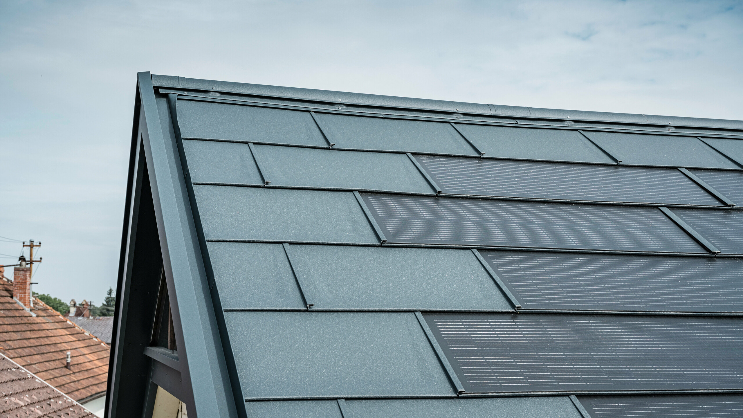 Vista dettagliata del tetto di una casa dotata dell'innovativa tegola PREFA Solar. Le tegole con celle fotovoltaiche integrate si presentano in un elegante colore antracite. La superficie omogenea si integra perfettamente nel tetto, creando un aspetto moderno e pulito. L'innovativo sistema di copertura garantisce un utilizzo efficiente dell'energia senza compromettere l'estetica.