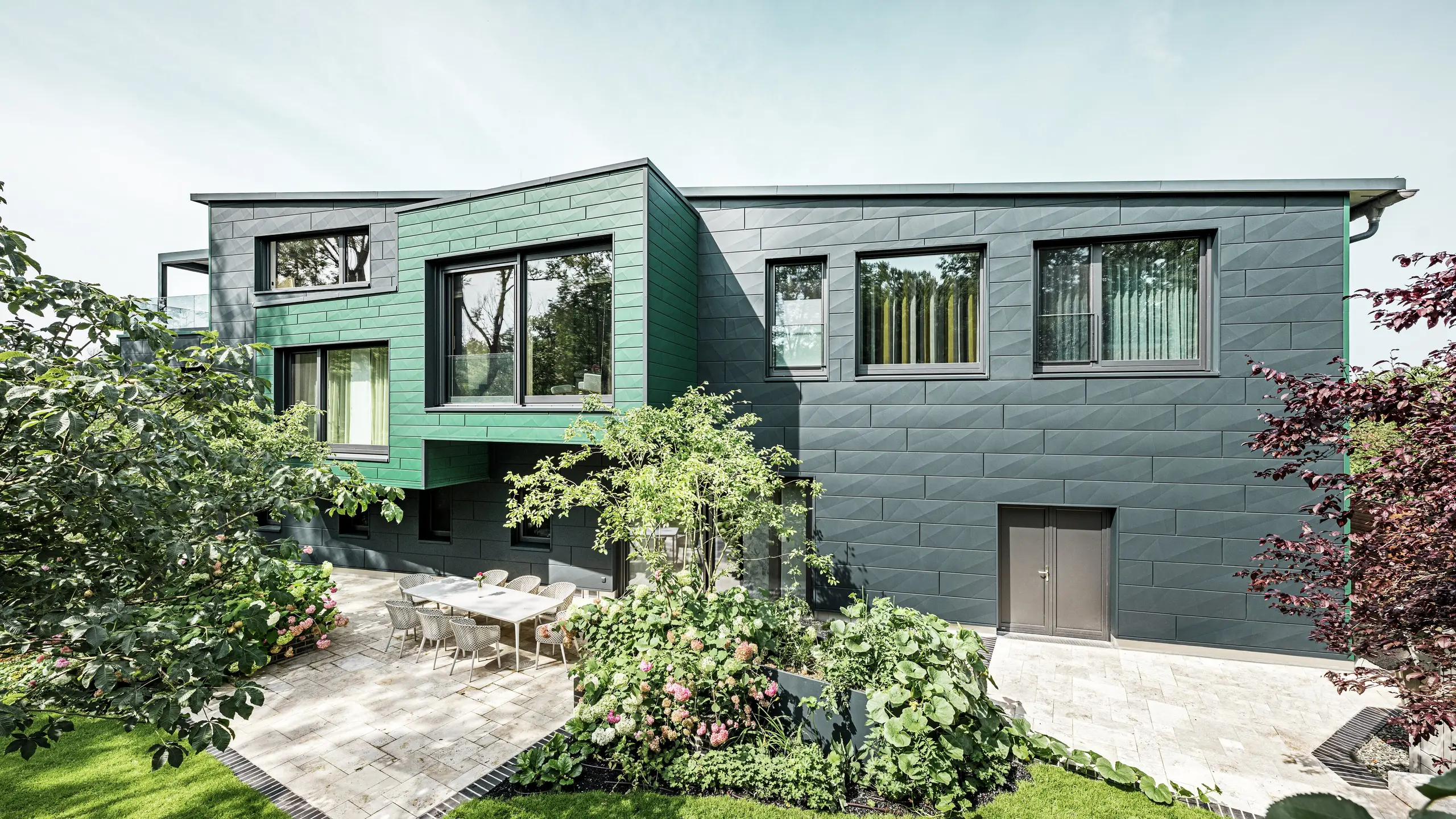La casa unifamiliare a Türkheim colpisce per la sua facciata straordinaria, rivestita con pannelli PREFA Siding X nei colori antracite e verde muschio. Le linee pulite e la scelta cromatica decisa dei pannelli in alluminio conferiscono all'edificio un aspetto moderno ed elegante. In primo piano si vede l'ampio giardino con un'elegante terrazza e un tavolo da pranzo con sedie moderne. Aiuole colorate e piante rigogliose creano un vivace contrasto verde con la robusta facciata. La facciata in alluminio, resistente e di facile manutenzione, protegge l'edificio e garantisce una bellezza senza tempo.