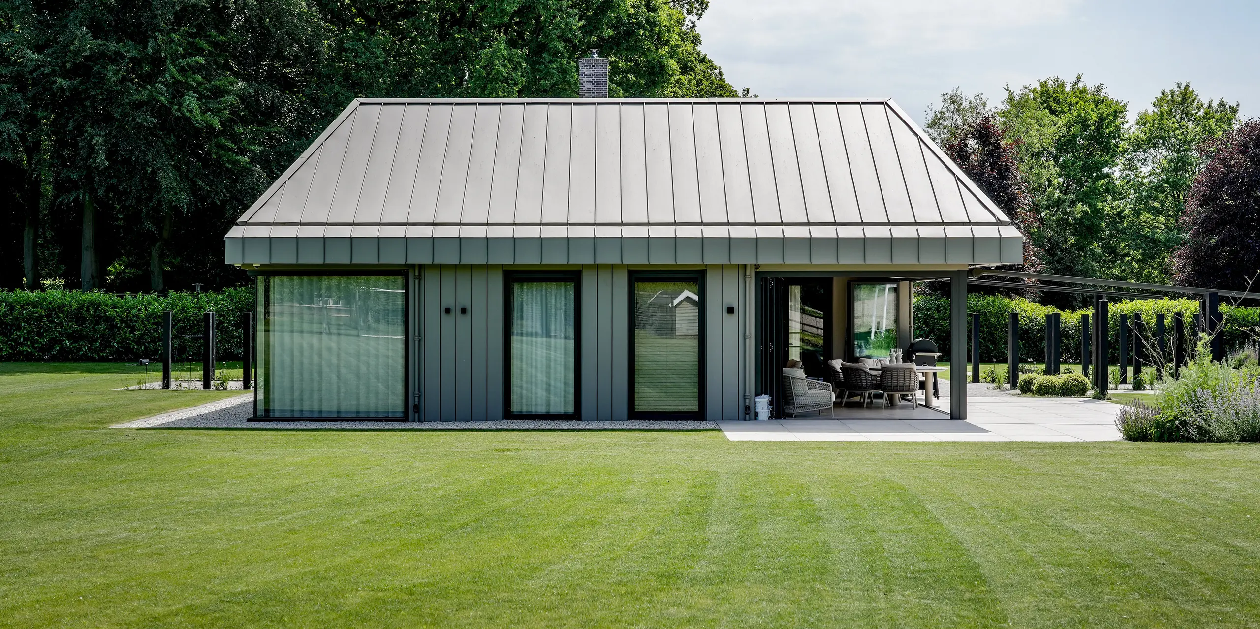 Frontalansicht des modernen Erweiterungsbaus in Putten mit durchgängiger PREFALZ Aluminiumverkleidung für Dach und Fassade. Die präzisen Stehfalze, die klar definierte Dachform und die großzügigen Glasflächen schaffen eine elegante, zeitlose Architektur. PREFA Aluminium überzeugt durch langlebige Qualität, hohe Witterungsbeständigkeit und eine harmonische Einbindung in die weitläufige Gartenlandschaft.