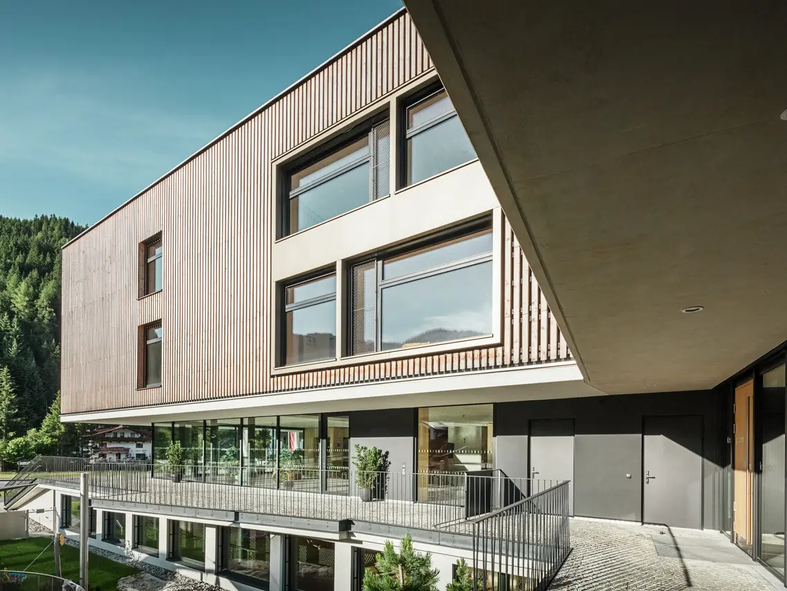  Un edificio a più piani dall'architettura moderna con una combinazione di legno e facciata in alluminio, in un contesto alpino con colline boscose sullo sfondo. L'edificio è dotato di ampie finestre che lasciano entrare molta luce e di una terrazza coperta che sottolinea l'ingresso.