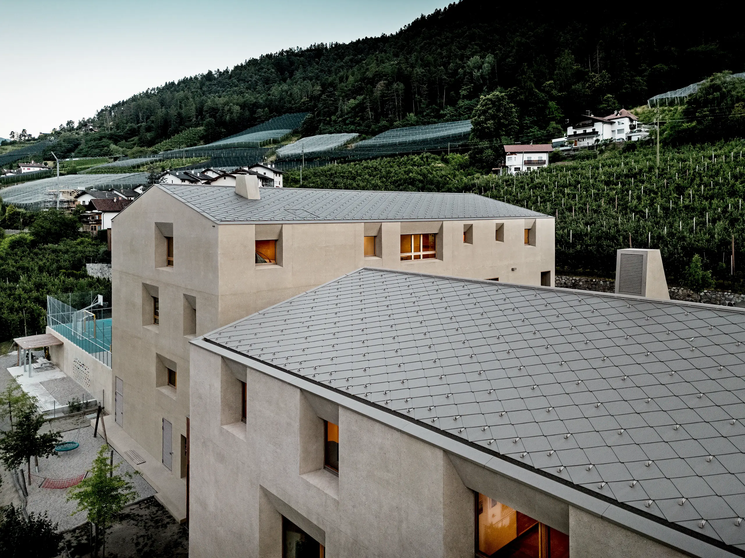 Da una prospettiva a volo d'uccello, il centro educativo di Riffian in Alto Adige mostra le sue caratteristiche scaglie 44 × 44 PREFA in P.10 bronze. La copertura in alluminio, realizzata con precisione, si estende armoniosamente sull'asilo e sulla scuola elementare, garantendo protezione dalle intemperie e lunga durata, e inserisce in modo suggestivo l'architettura moderna tra i frutteti e il versante della montagna.