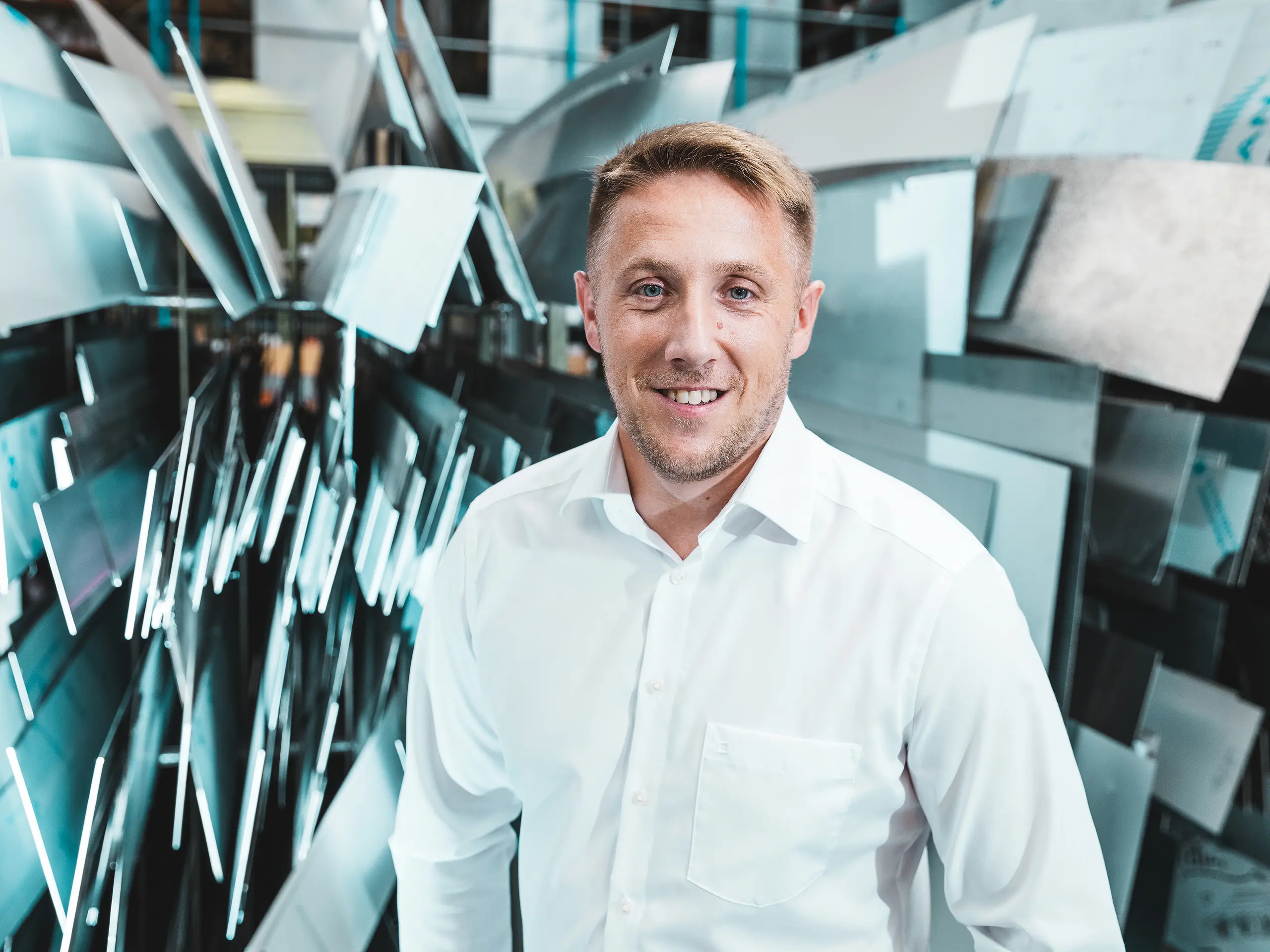 Selbstbewusst und freundlich steht Projektleiter Marcel Schmidt von der Spenglerei ASA Schüßler vor einer eindrucksvollen Wand aus unterschiedlich geformten Aluminiumelementen. Die dynamisch arrangierten Metallplatten spiegeln das Licht effektvoll wider und symbolisieren Innovation, Präzision und Qualität in der Metallverarbeitung. Der Mann im weißen Hemd verkörpert Kompetenz und moderne Fertigungsexzellenz. Im Hintergrund wird die vielfältige Verarbeitungsmöglichkeit von Aluminium sichtbar – von Dach- und Fassadensystemen bis hin zu maßgefertigten Sonderlösungen. 