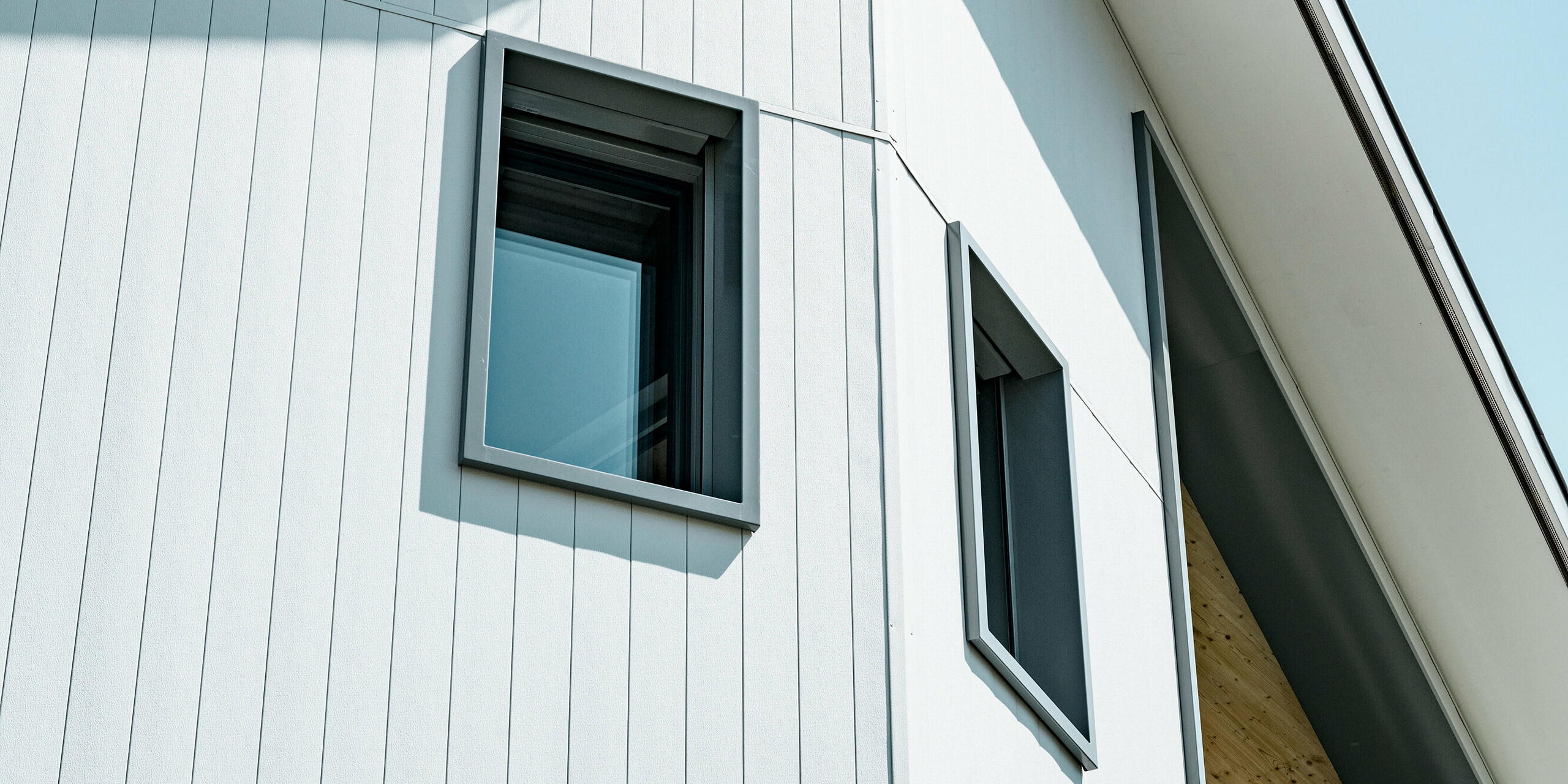 Detailaufnahme der klaren und modernen PREFA Siding Aluminiumfassade in P.10 Prefaweiß an einem Mehrfamilienhaus in Weissbad, Schweiz. Die präzise vertikale Ausrichtung der Sidings bildet eine ansprechende Textur und sorgt für ein dynamisches architektonisches Element. Die tief eingelassenen Fenster mit dunklen Rahmen kontrastieren effektvoll gegen die helle Fassade, betonen die geradlinige Ästhetik des Gebäudes und spiegeln die hohe Qualität der PREFA Aluminiumprodukte wider.