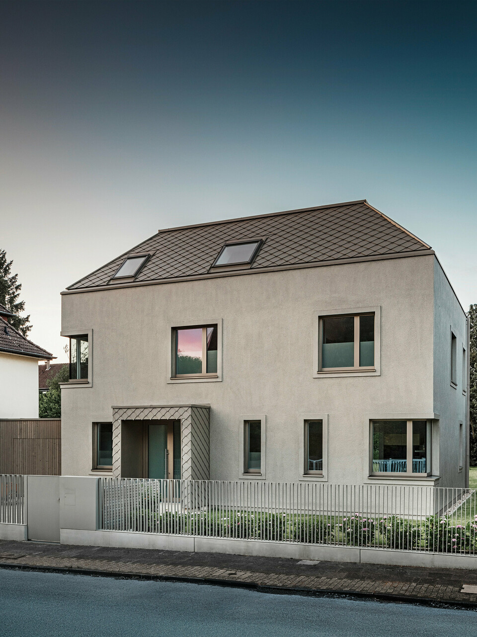 Una moderna casa indipendente con tetto a capanna e rivestimento di facciata e tetto in alluminio color bronzo di PREFA. La casa si trova in un tranquillo quartiere residenziale, circondato da giardini ben curati e da altre case unifamiliari. In primo piano si nota una bassa recinzione che separa la proprietà dalla strada. L'architettura della casa è caratterizzata da linee pulite e ampie finestre che lasciano entrare molta luce naturale all'interno. 