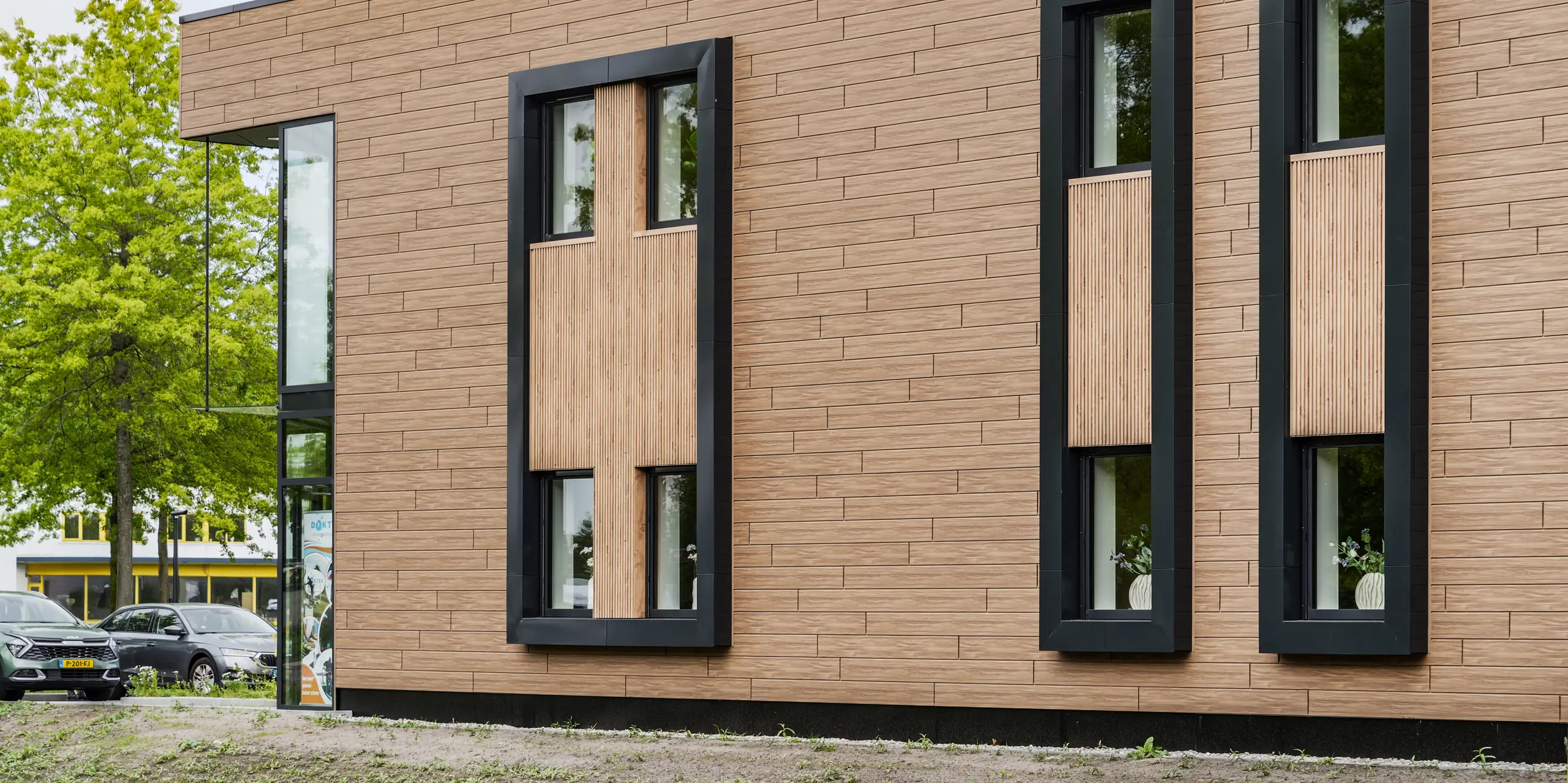 Das moderne Firmengebäude der Dokter Schoonmaakorganisatie in Wolvega beeindruckt mit seiner klaren, kubischen Architektur und markanten Fensterbändern. Die walnussbraune PREFA Fassade mit Sidings in authentischer Holzoptik verleiht dem Baukörper eine warme und hochwertige Ausstrahlung. Schwarze Rahmenelemente, großzügige Verglasungen und präzise Linienführungen schaffen ein spannendes Zusammenspiel aus Eleganz und moderner Fassadengestaltung.